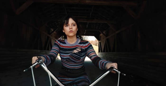 Jenna Ortega rides a bike through a covered bridge, wearing a striped sweater. She looks ahead with a determined expression