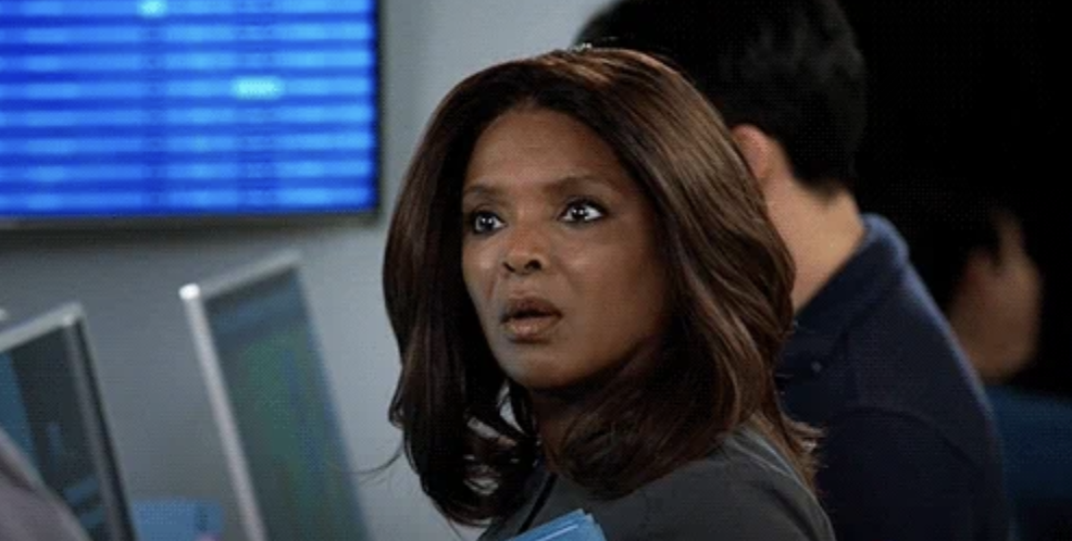 A woman with long hair looks surprised, standing in front of computer screens. Background shows an office setting with multiple people