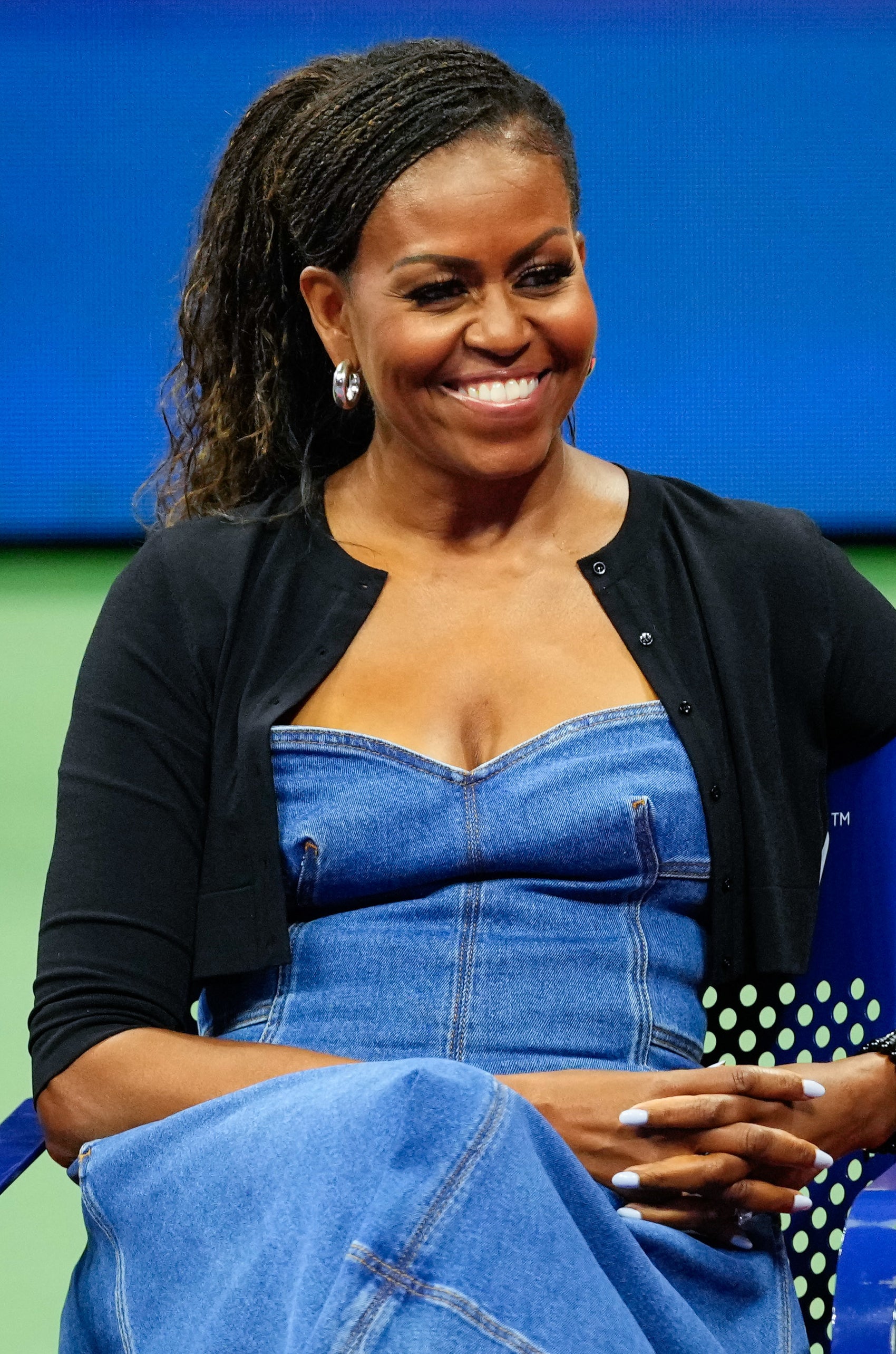 Michelle Obama sits smiling while wearing a chic denim dress and black cardigan