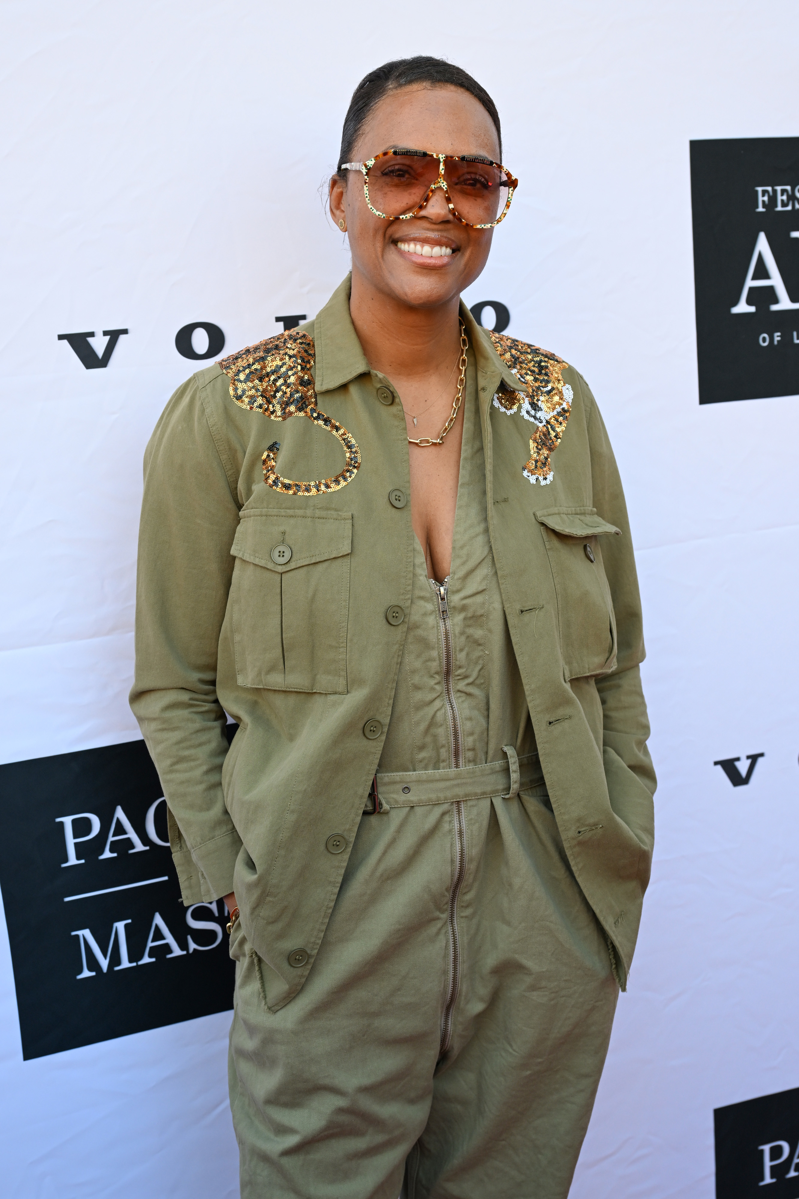 Deon Cole on the red carpet wearing an olive green jumpsuit with sequin embellishments, sunglasses, and a gold chain necklace