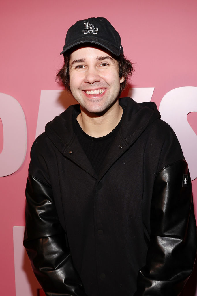 David Dobrik smiling while wearing a casual black jacket and black baseball cap on a red carpet