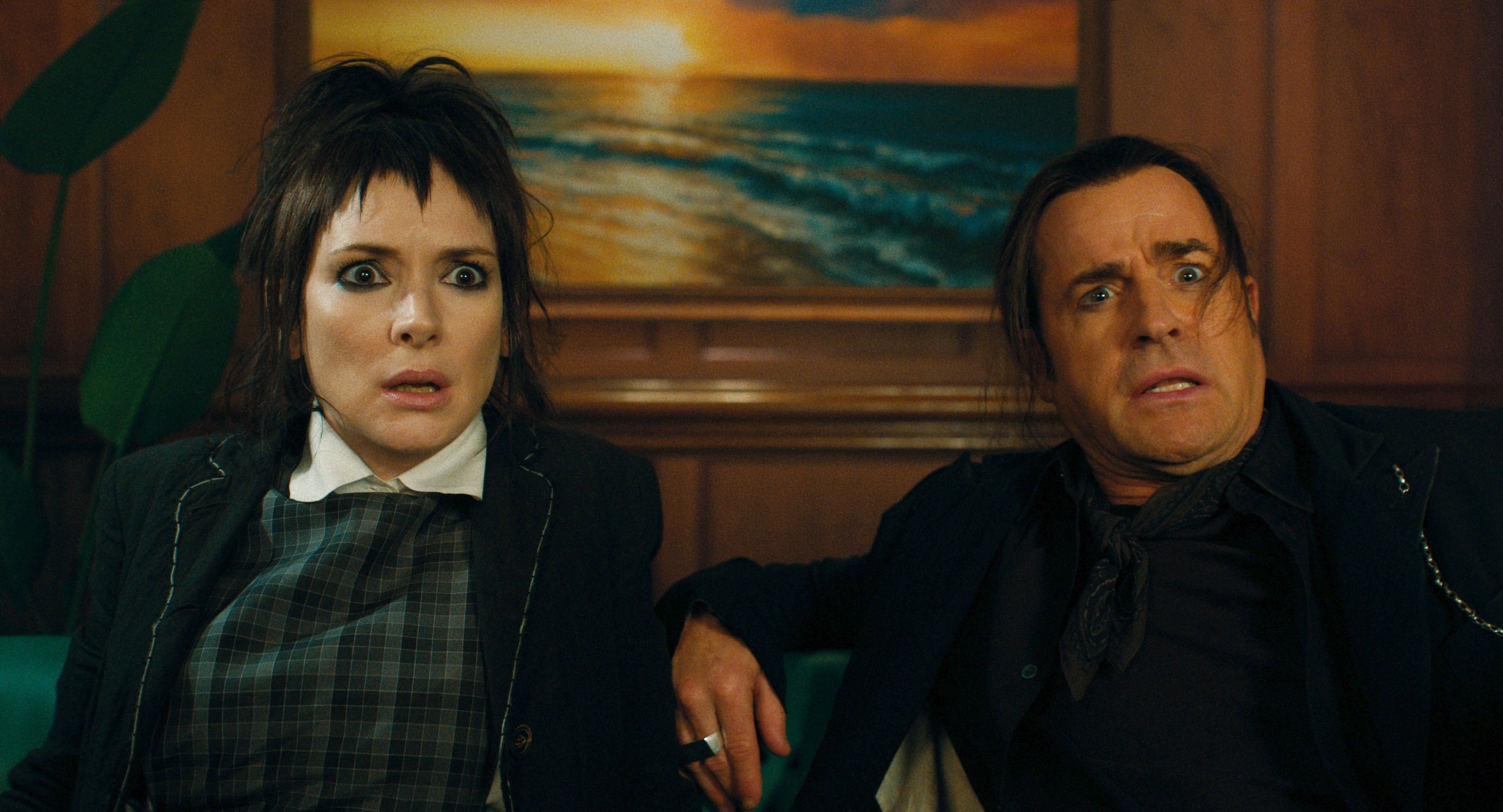 Winona Ryder and Justin Theroux look surprised sitting on a couch with a painting of a beach sunset behind them
