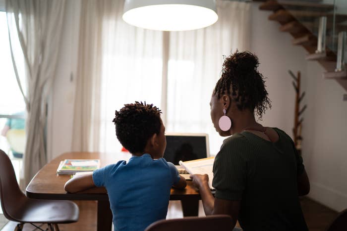 A woman and a child sit at a table with a laptop and papers, engaging in a discussion or study session in a home setting
