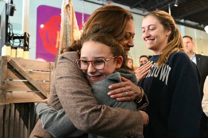 Kamala Harris hugs a young girl wearing glasses and a grey sweatshirt. Another young woman in a navy sweatshirt looks on with a smile