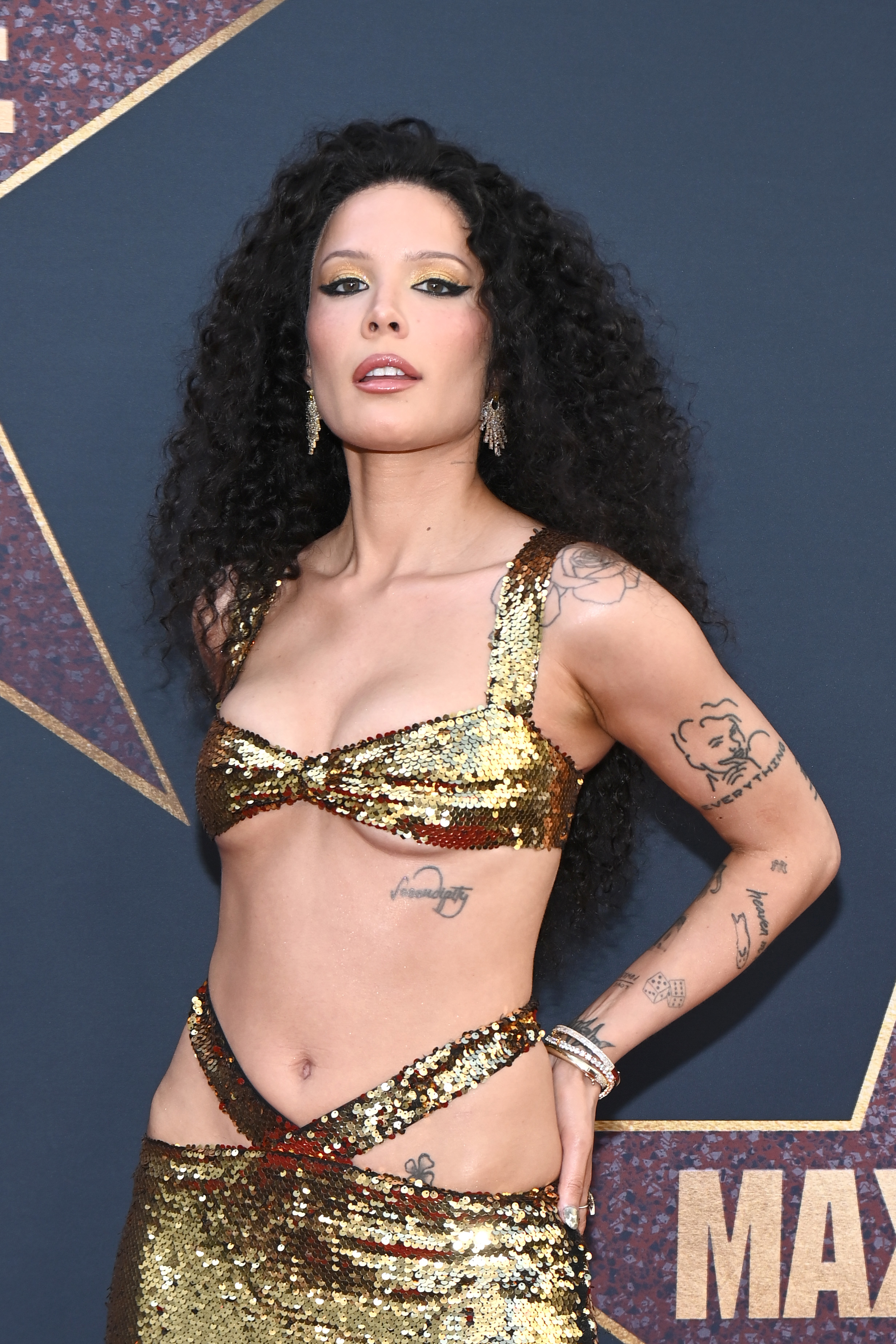Halsey poses on the red carpet in a gold sequin cut-out dress, showcasing tattoos on her arms and torso