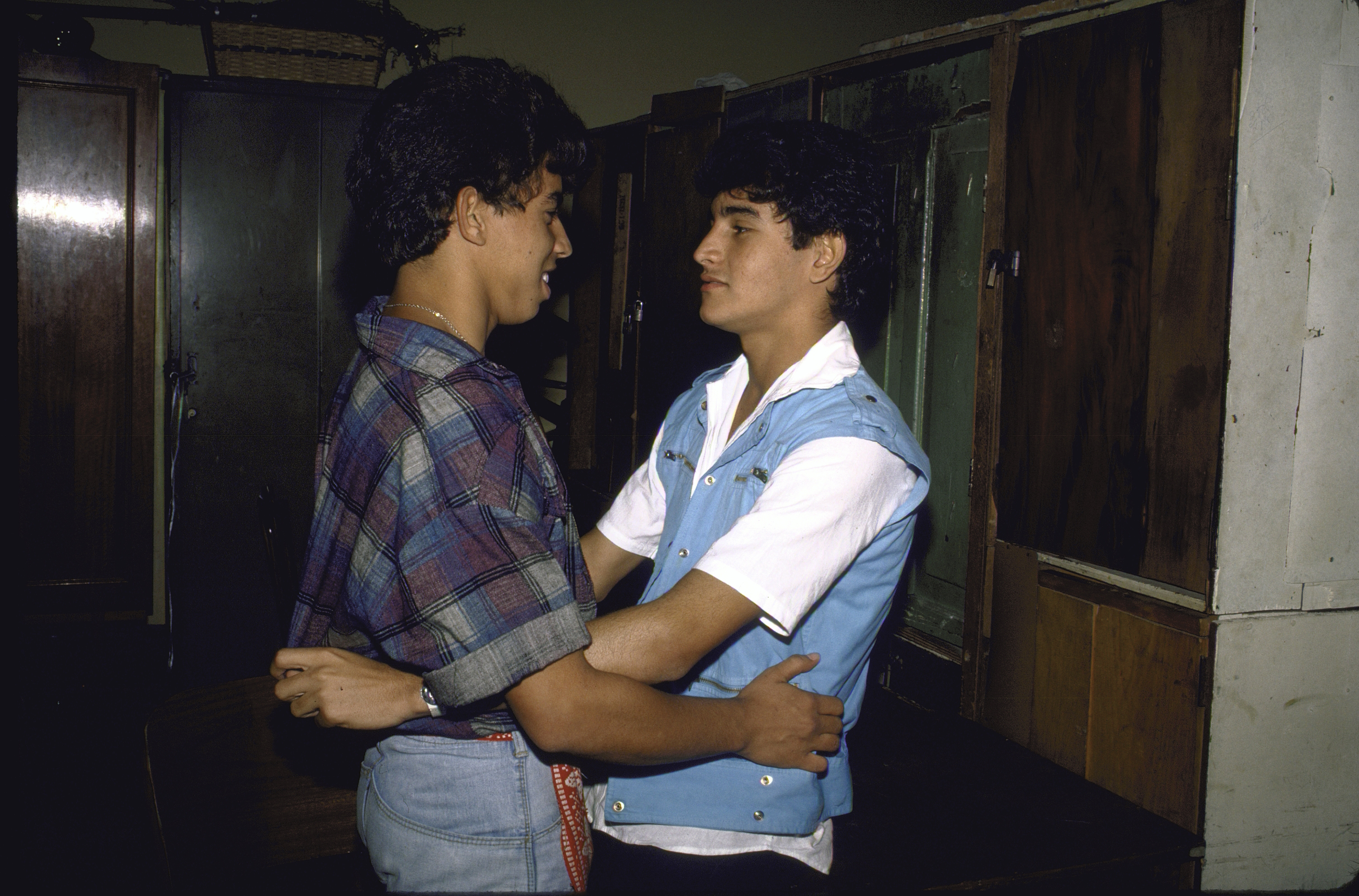 Two young men, one in a plaid shirt and the other in a white shirt with a blue vest, hold each other and gaze into each other's eyes in a room with wooden furniture