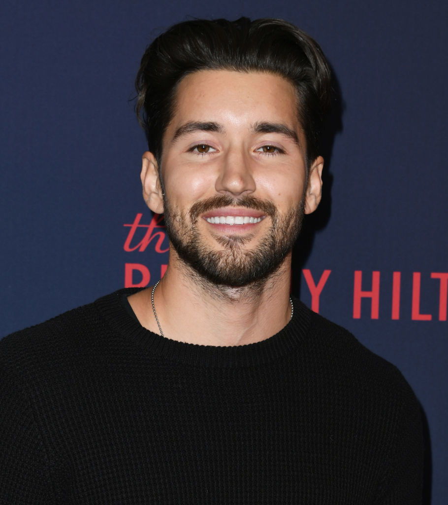 A man with a beard and short dark hair smiles while wearing a black sweater, standing in front of a backdrop for The Beverly Hilton. Names are unknown