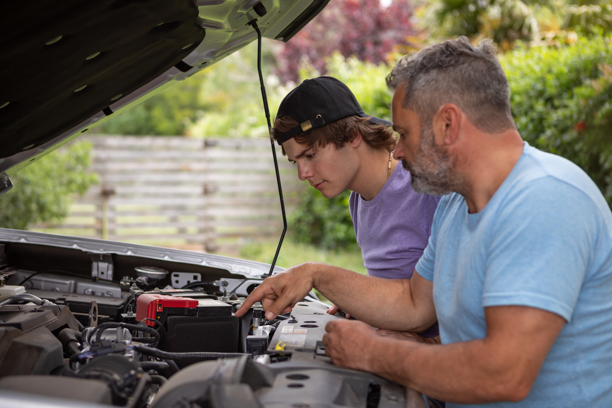 Two people, one younger wearing a backward cap, and one older with gray hair, are looking under the hood of a car and discussing its engine