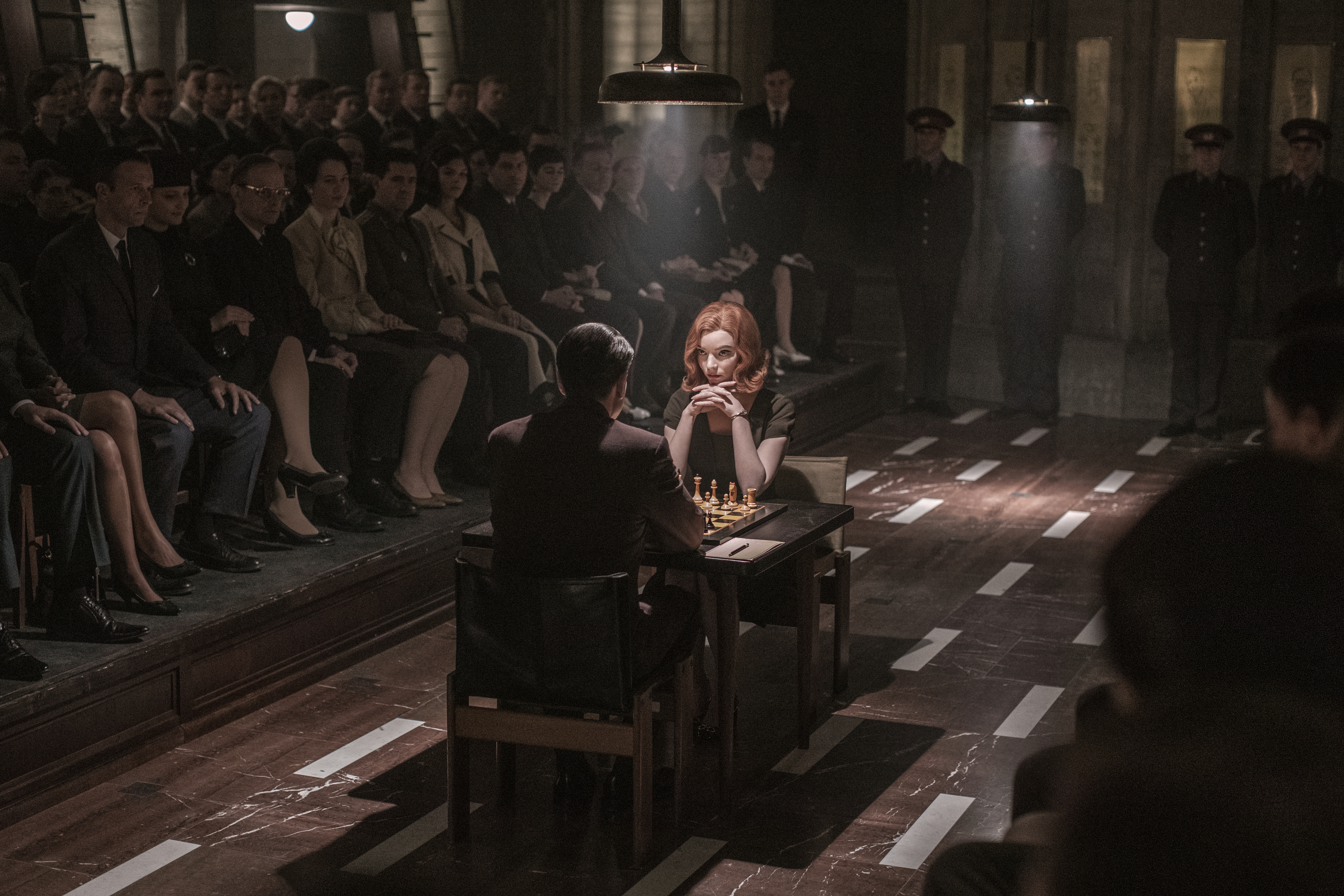 Beth Harmon plays a game of chess in a crowded room, intensely focused on her opponent, from a scene in "The Queen's Gambit."