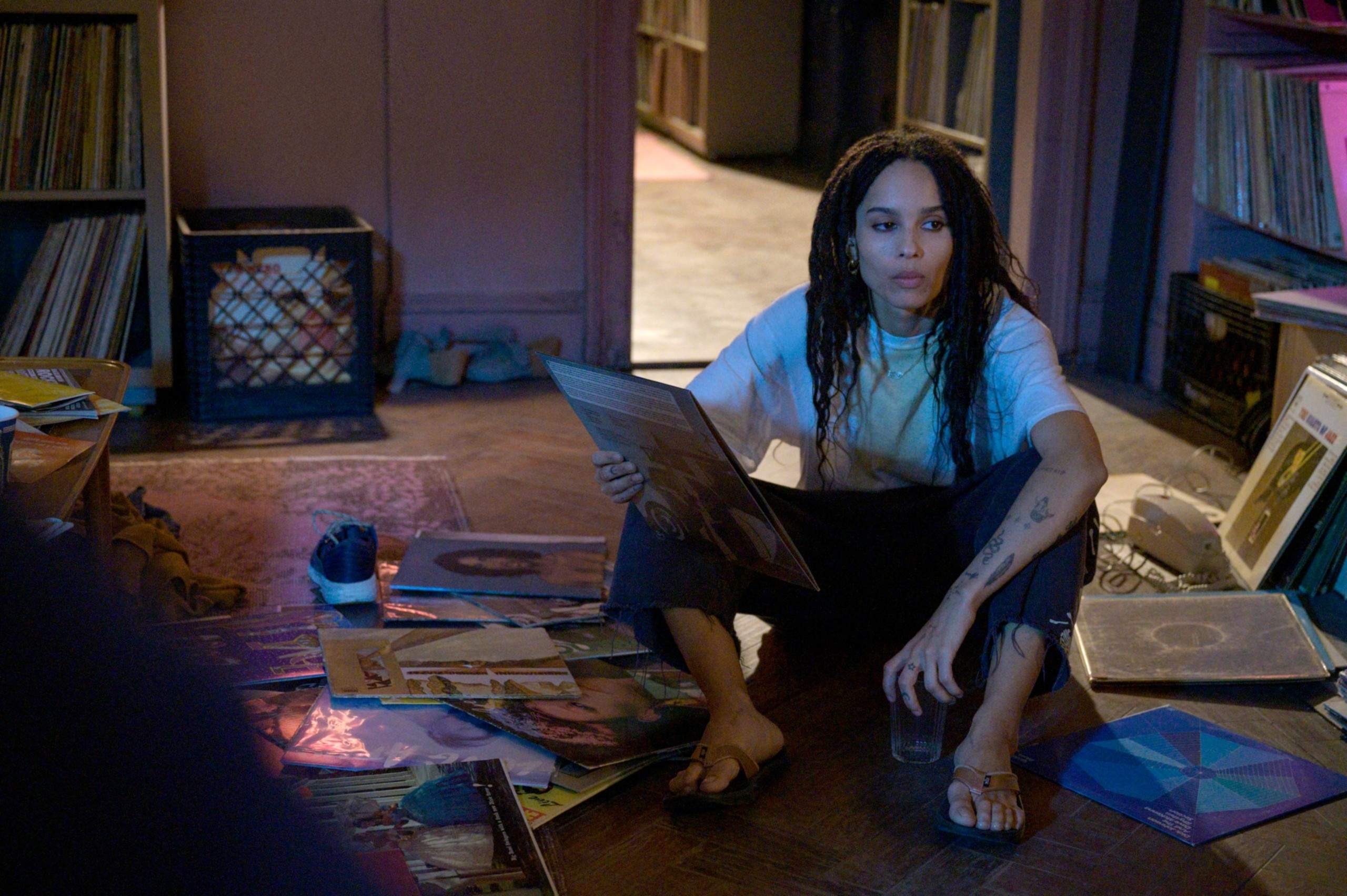 Zoë Kravitz sits on the floor among scattered vinyl records, wearing a casual white t-shirt and dark pants, appearing contemplative