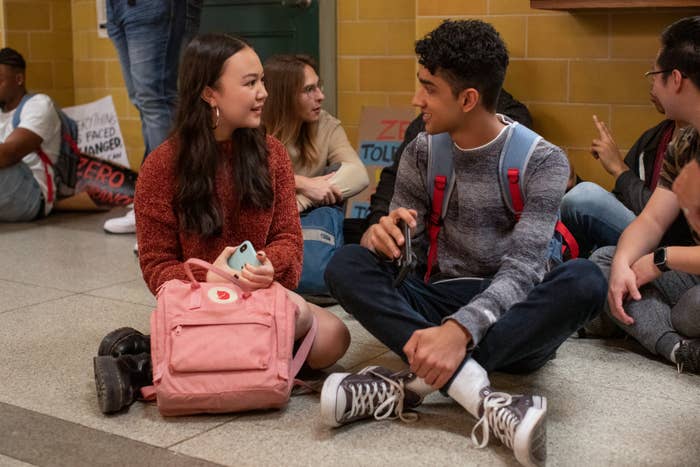 Anna Cathcart, seated with a pink backpack, and Jaren Lewison, in a gray sweater, talk in a school hallway setting with other students around