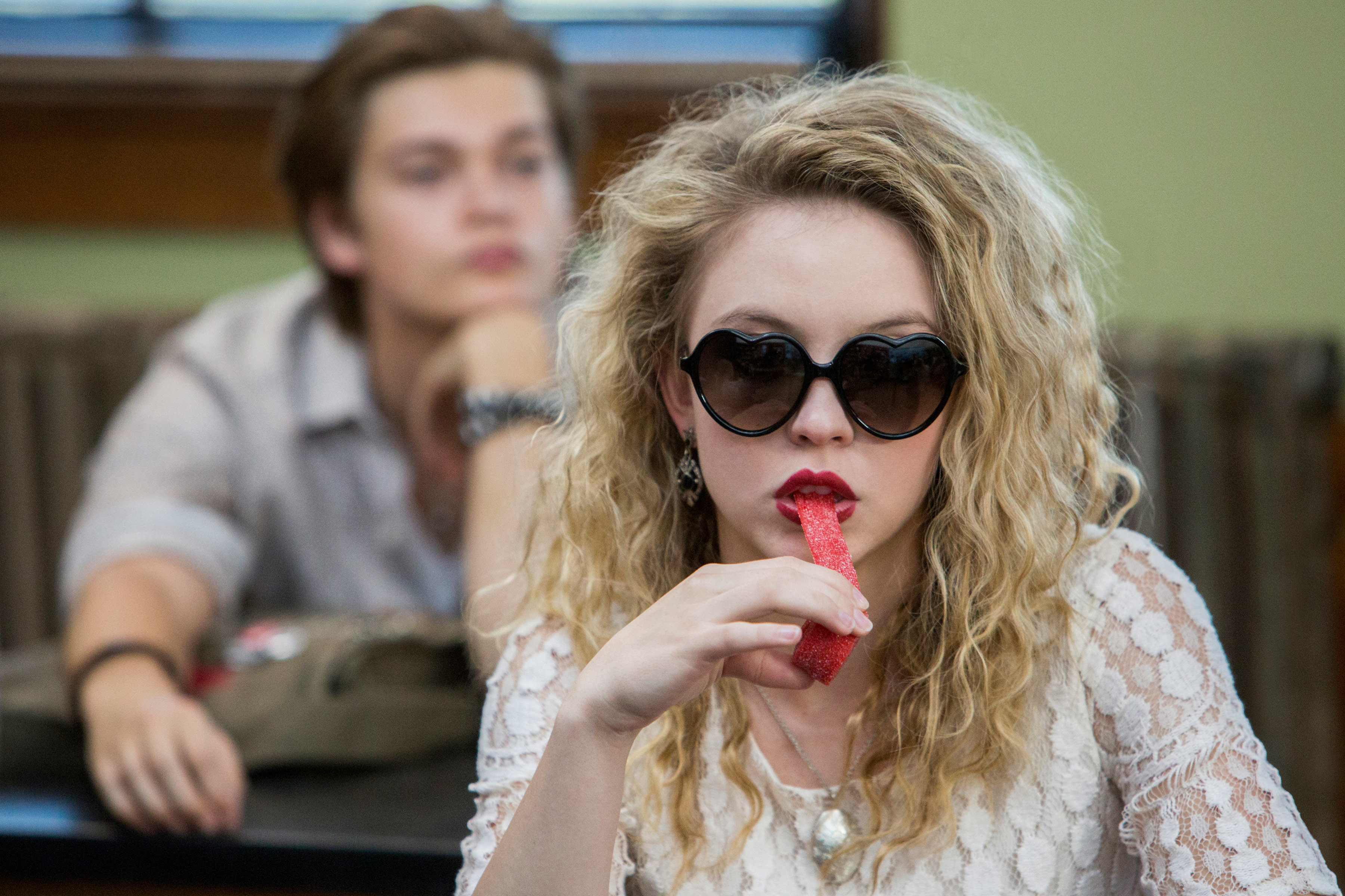 A character from the TV show "Everything Sucks!" wearing heart-shaped sunglasses and eating candy, with a blurred background of a classroom and another student