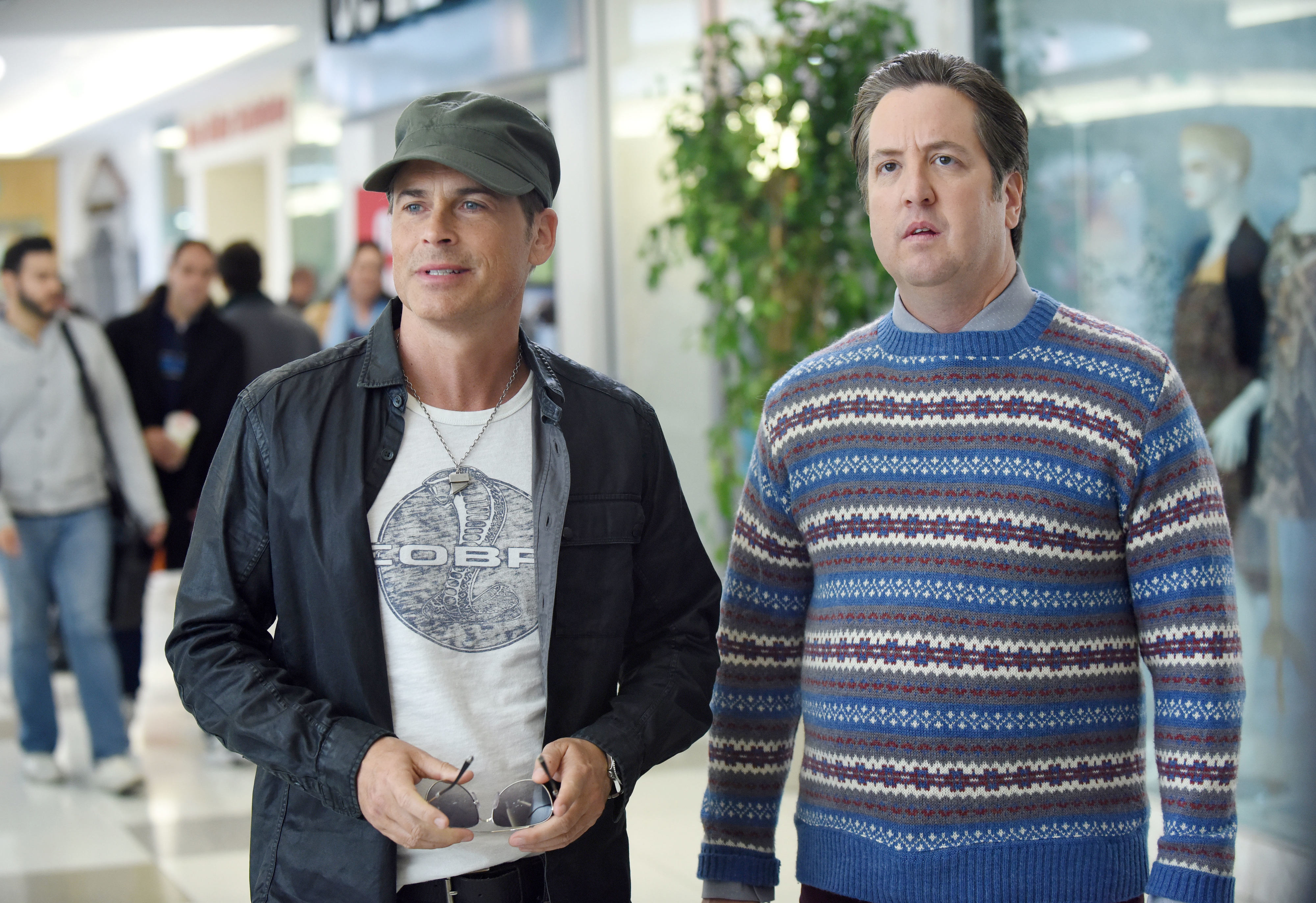 Rob Lowe, wearing a casual jacket and cap, stands beside Nate Torrence in a patterned sweater in a brightly lit shopping mall