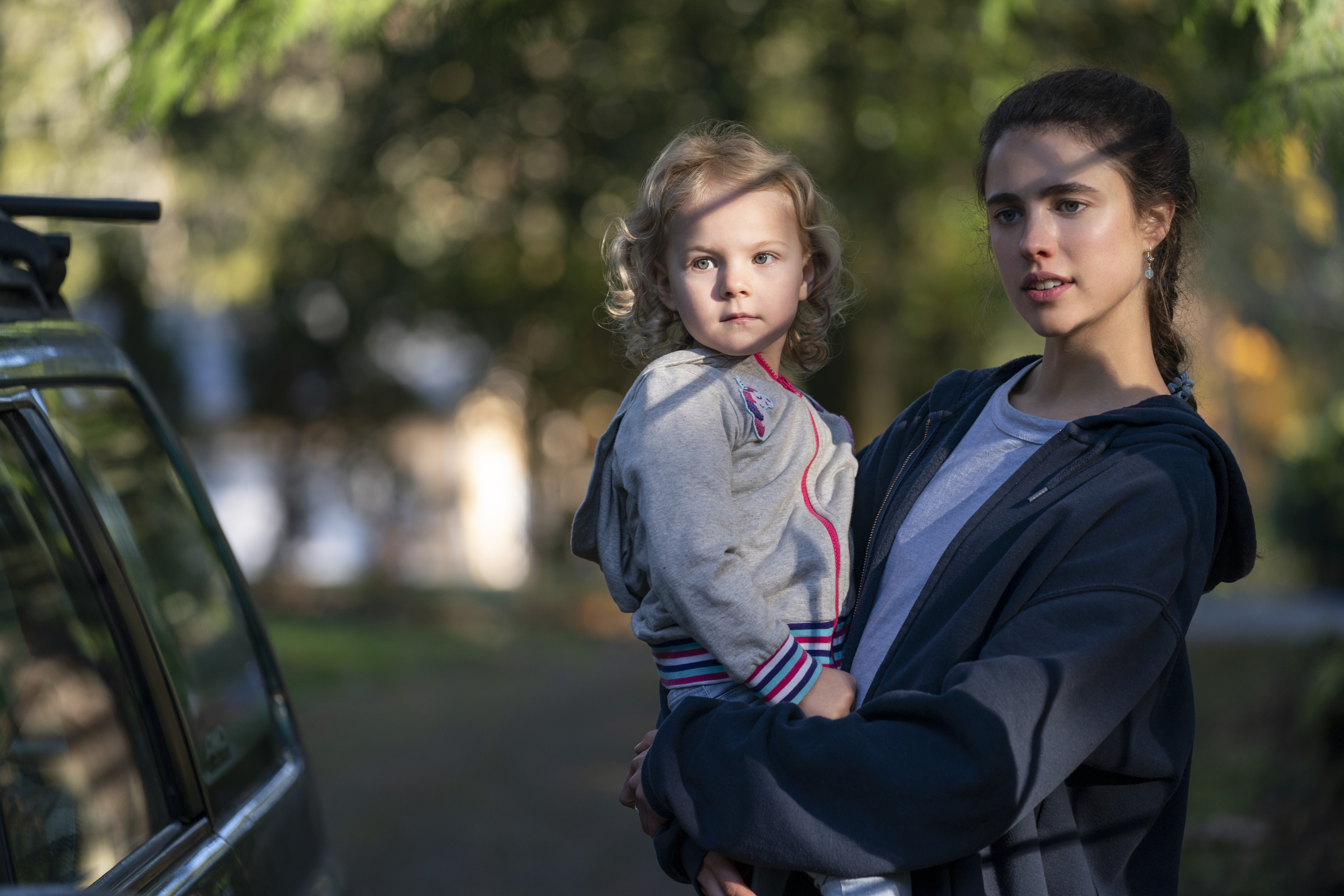 Margaret Qualley holds a young child while standing next to a car in a scenic outdoor setting, likely from a TV show or movie scene