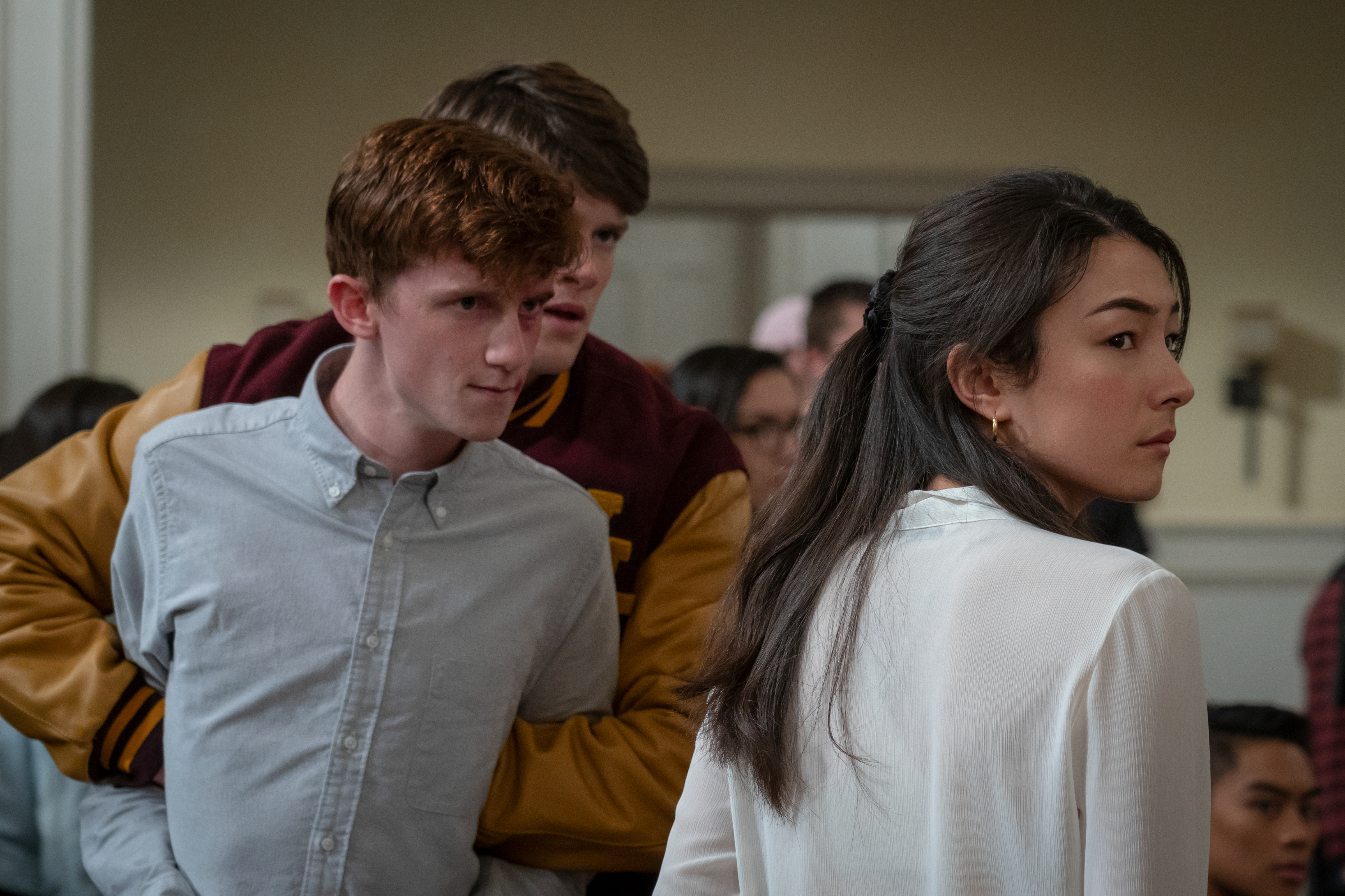 A man in a light shirt speaks with a serious expression, alongside another male in a varsity jacket, and a woman with a concerned look in a white blouse