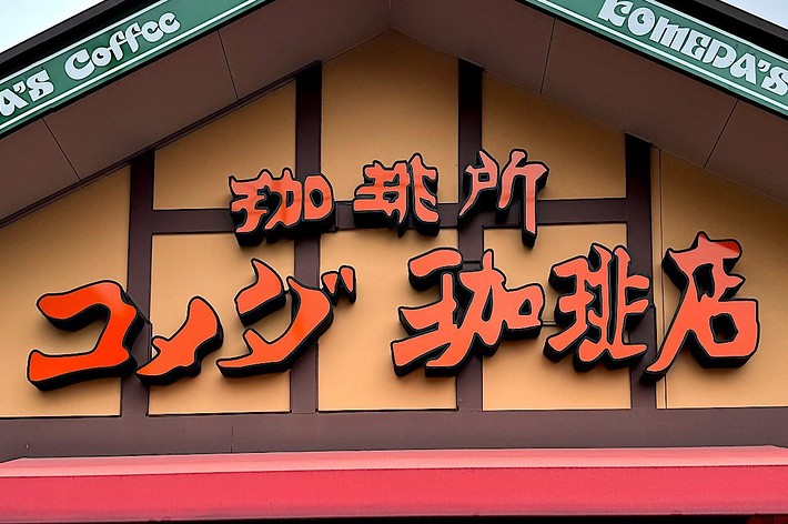The image shows the exterior of a cafe with a sign that reads “Komeda’s Coffee” in both English and Japanese. The building has a red awning