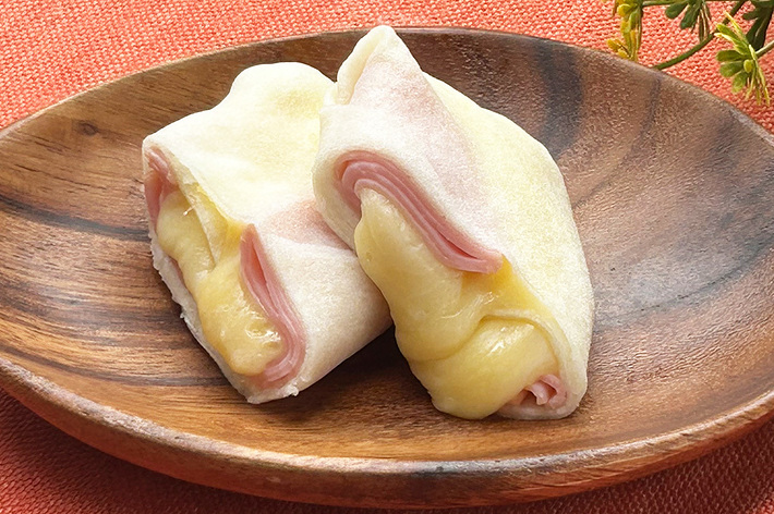 木製の皿に乗った2つのハムとチーズを包んだパン生地、背景には花が飾られている。