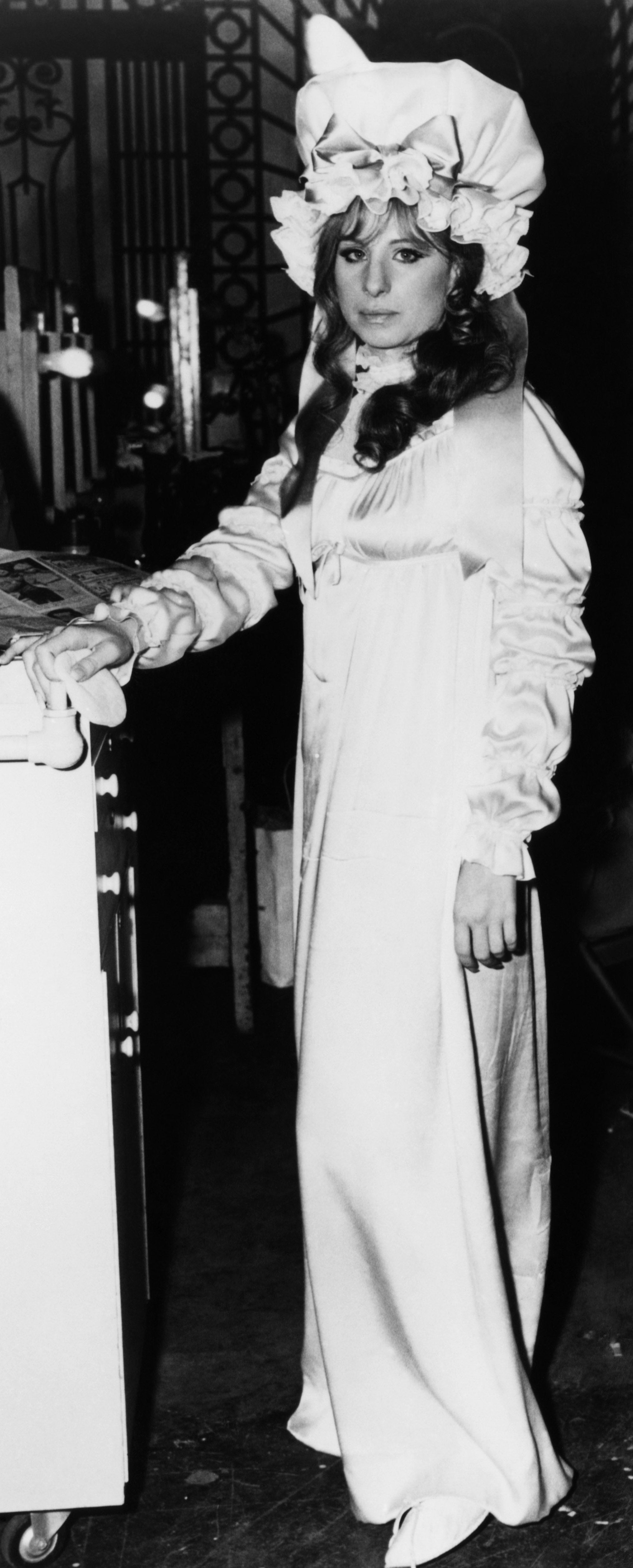 Barbra Streisand stands beside a sketch of herself in an elegant, long-sleeved dress with puffed shoulders and a bonnet, matching the illustration