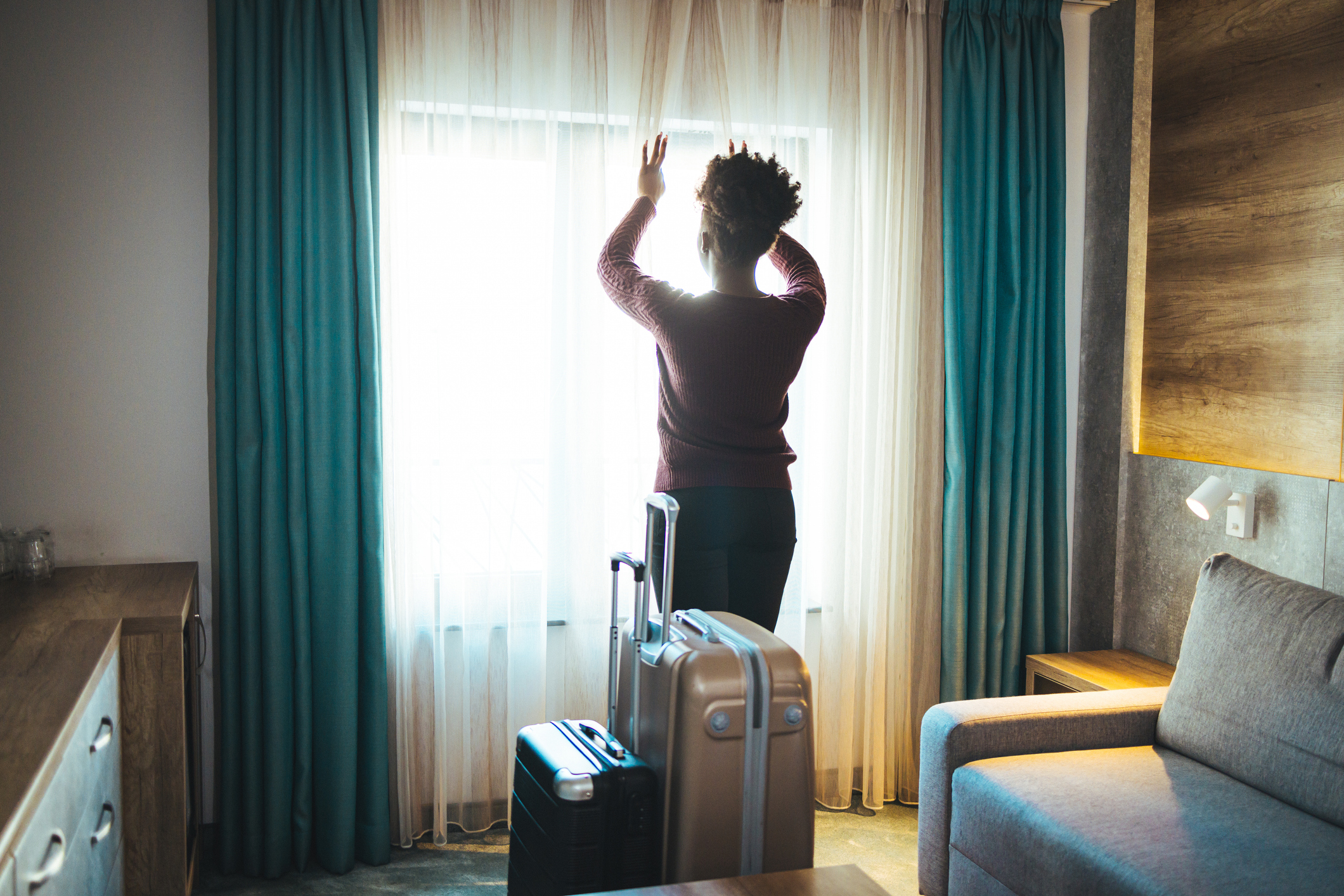 A woman looks out of a hotel window while holding the curtains. Two suitcases are next to her. The image is themed around travel and accommodation