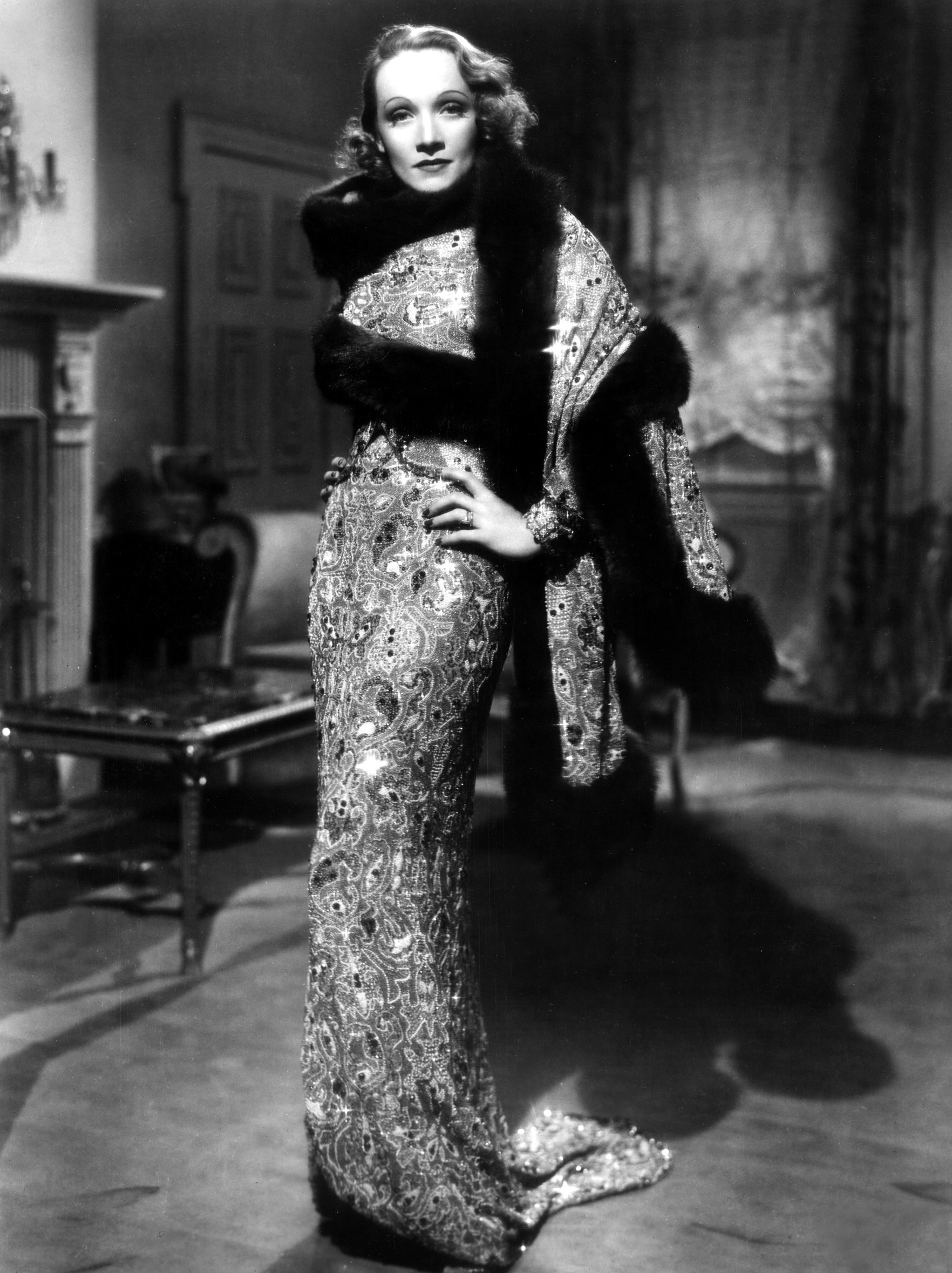 Marlene Dietrich in an elegant, form-fitting, beaded gown with a luxurious fur stole, posing gracefully indoors