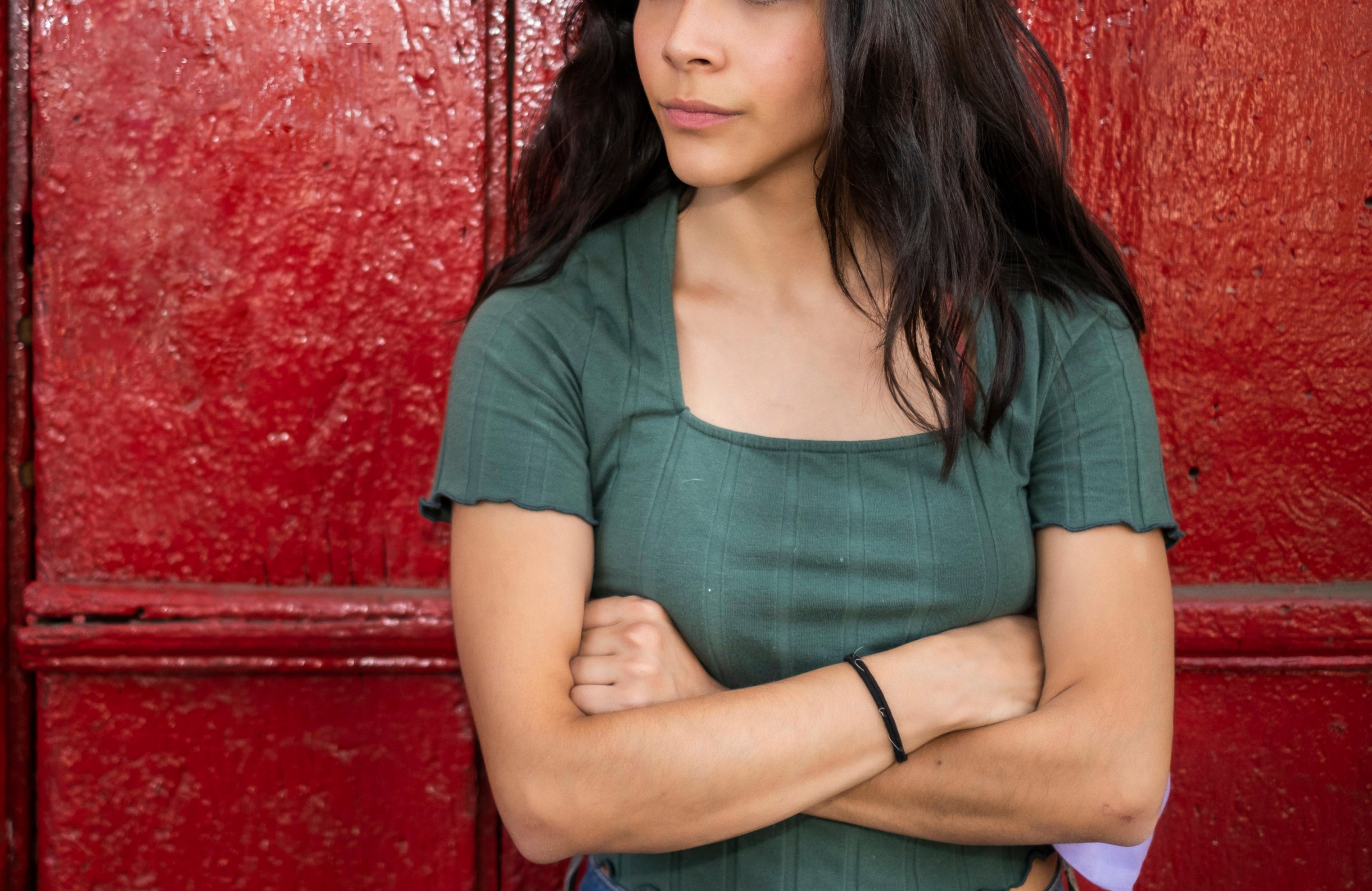 A woman with long hair in a short-sleeved top stands with arms crossed against a red wooden background, looking to the side