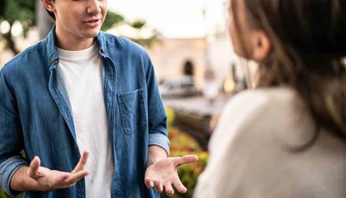 A young man in a casual shirt is talking to a woman outdoors near trees and buildings. Both are engaged in conversation.