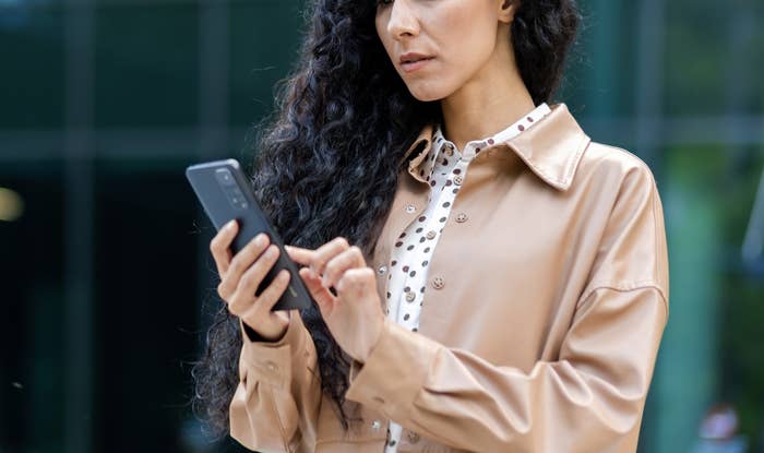 A woman with long curly hair, wearing a beige coat over a polka-dot blouse, looks intently at her smartphone outside a modern glass building