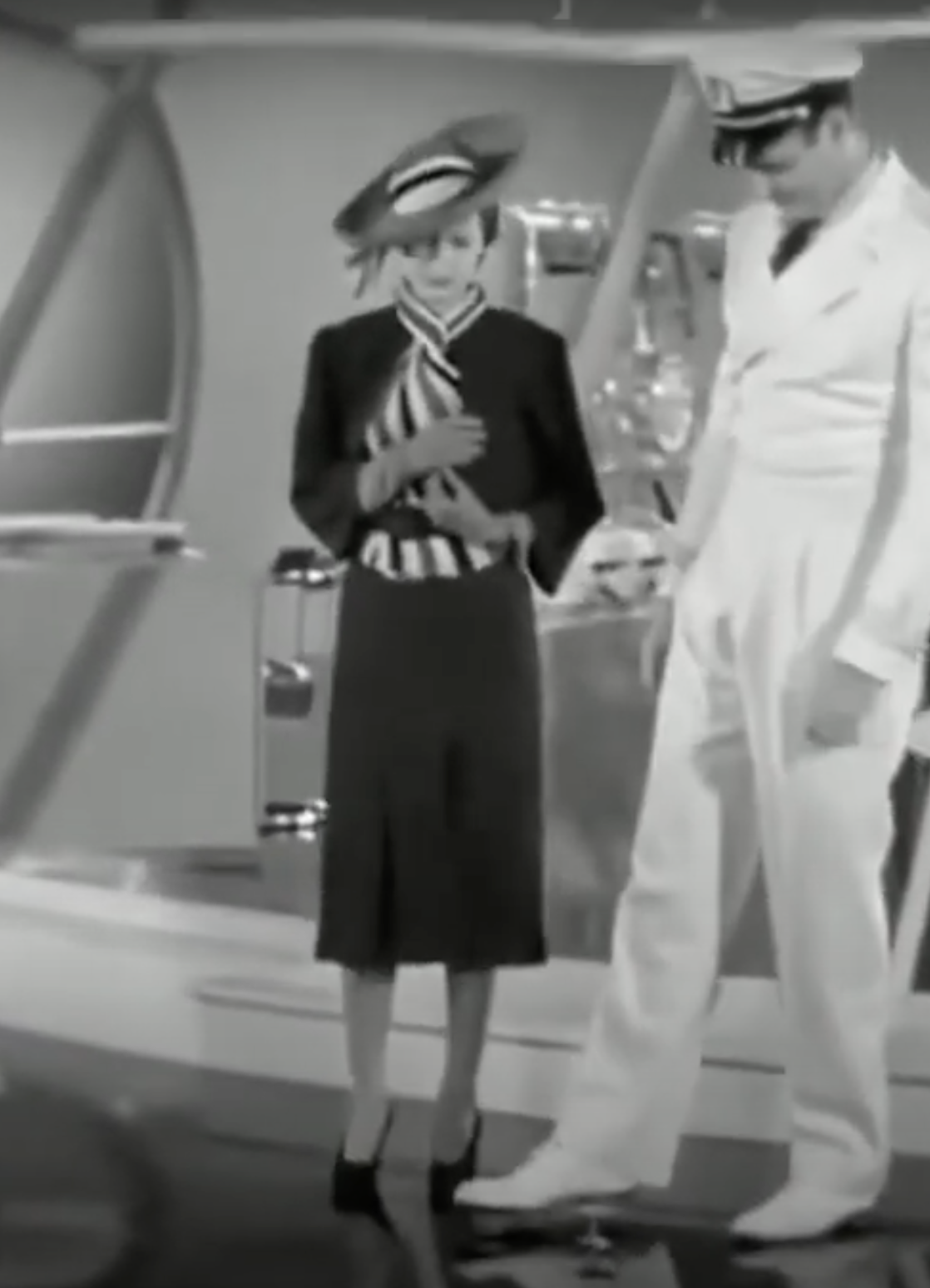 Dorothy Lamour wears a striped scarf and hat, next to someone is in a white naval uniform. They have focused expressions