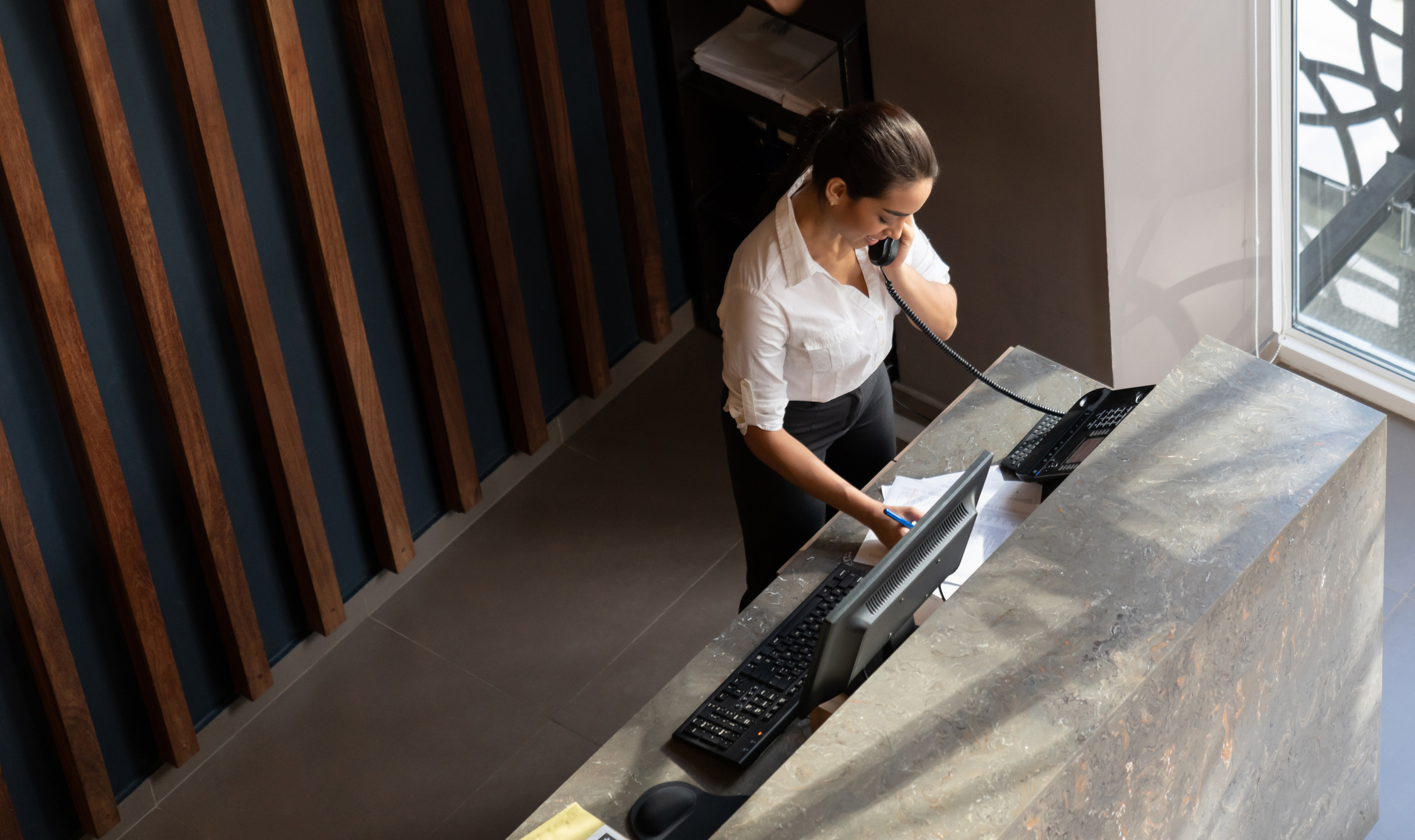 A receptionist speaks on the phone while writing down notes at a hotel front desk, ready to assist guests