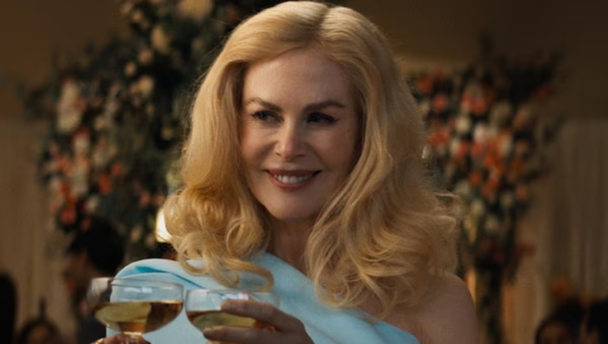 Nicole Kidman sonriendo, sosteniendo dos copas de vino blanco en una fiesta elegante con un fondo de flores decorativas