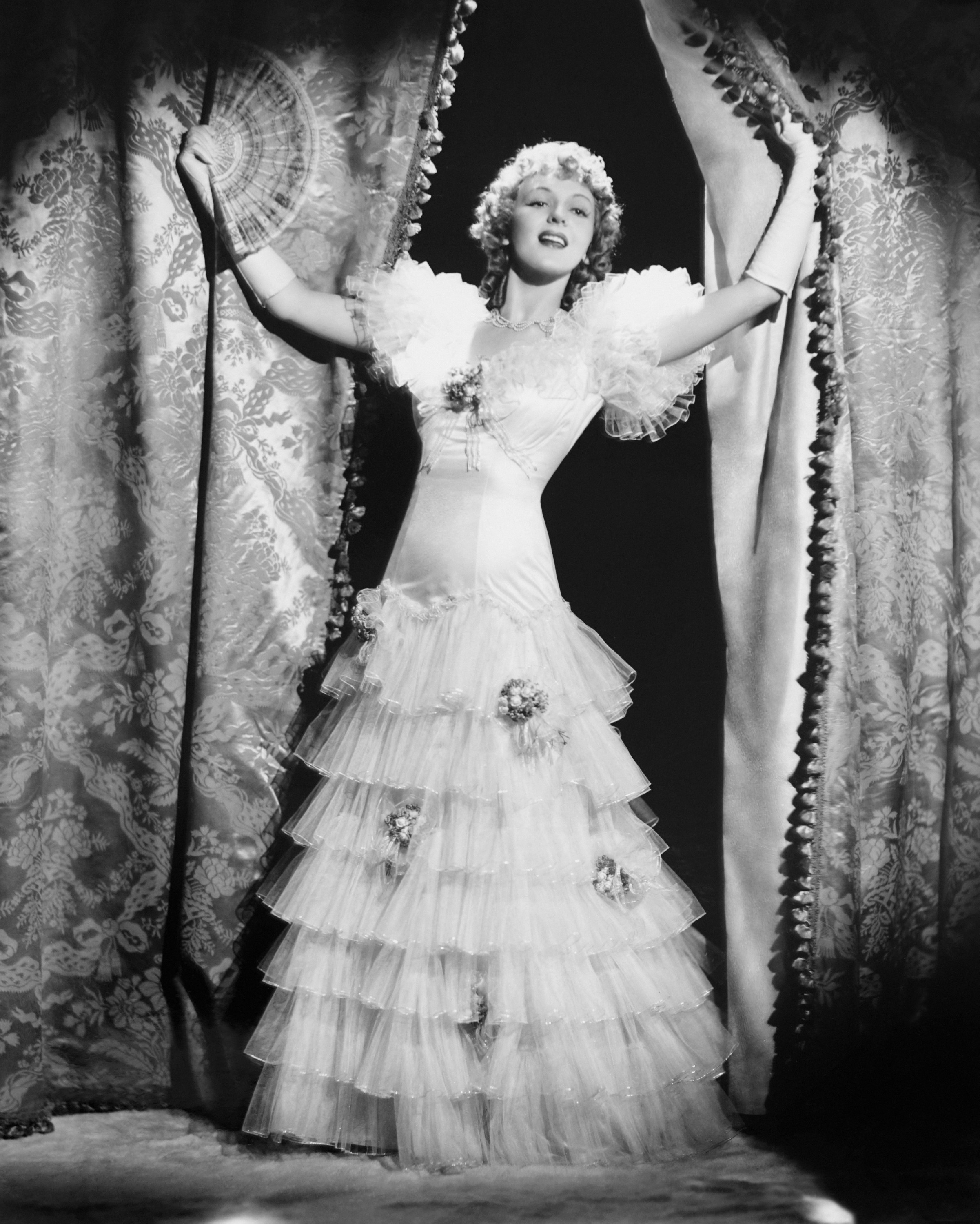 Woman in elaborate vintage dress with ruffled layers and flower details, holding open heavy curtains in a theatrical pose. I don't know who she is