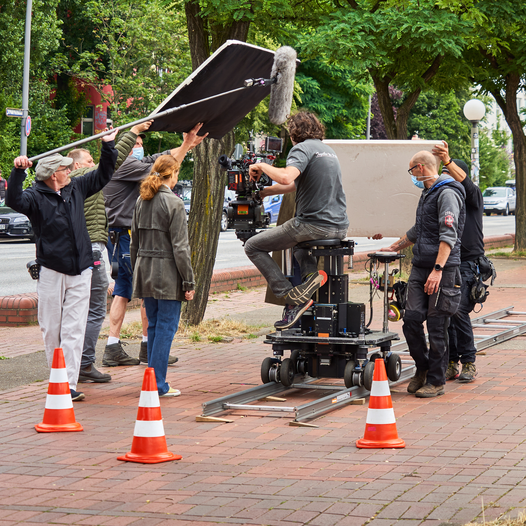 Film crew on a set with various equipment, including a camera on a dolly and lights. Trees and traffic in the background