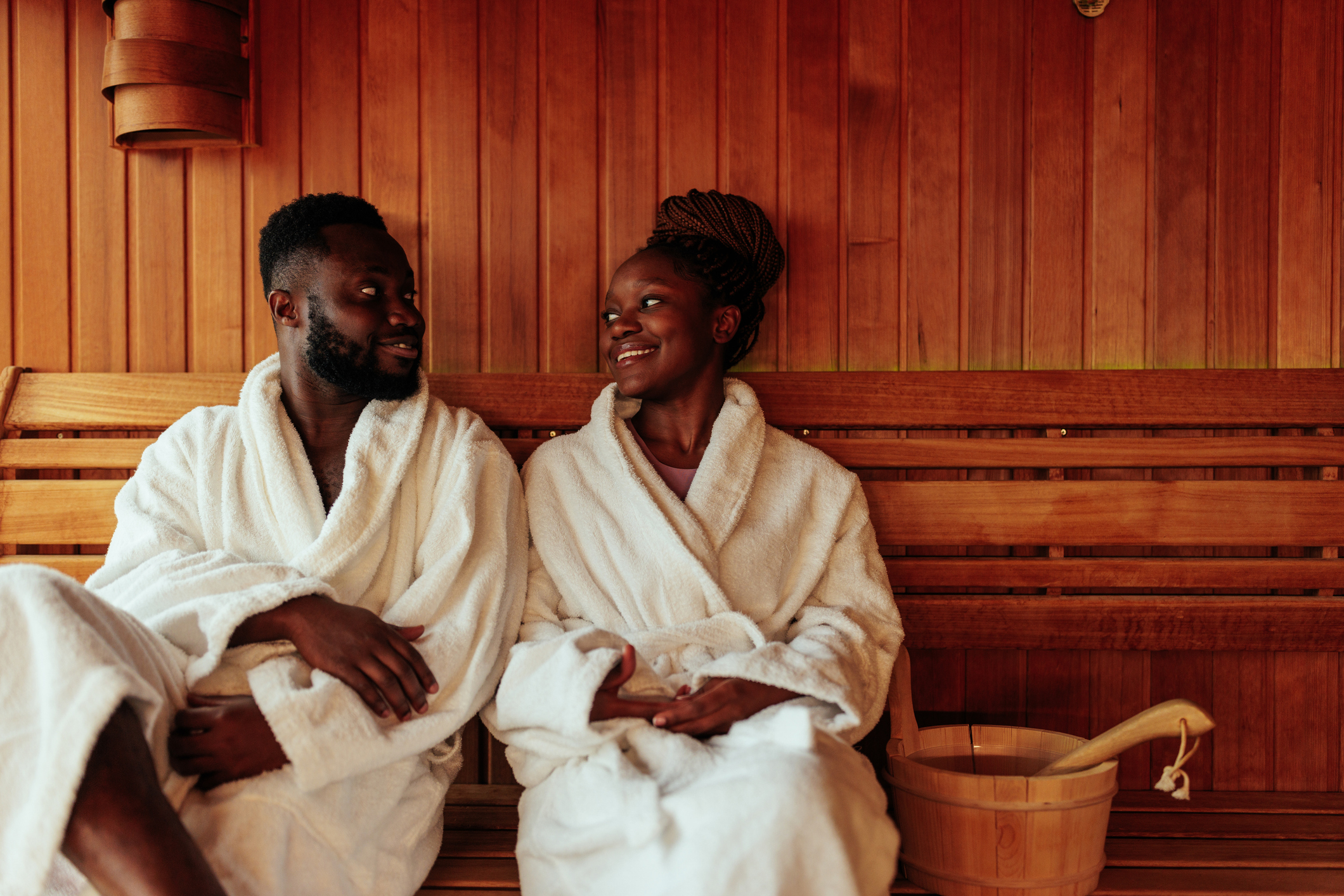 Two people in white robes relax and smile at each other inside a wooden sauna