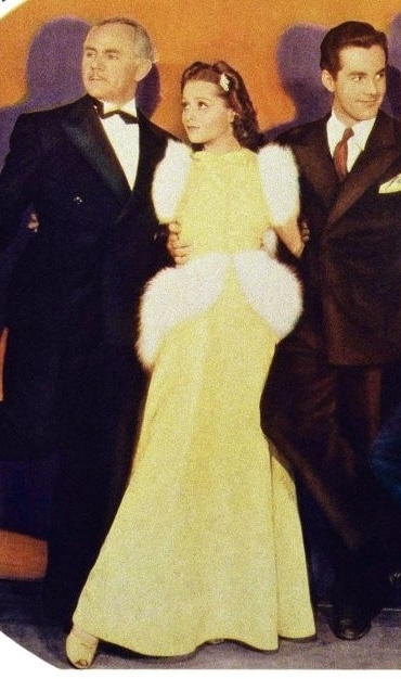 Three unidentified people stand together, dressed in formal attire. The person in the center wears a long dress with white fur accents. The men wear suits