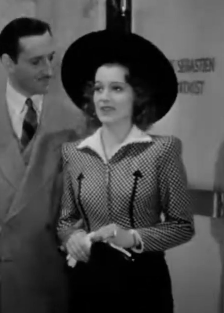 Two people in vintage attire, including a man in a suit and a woman wearing a large hat and a patterned dress with white accents, standing in front of an office door