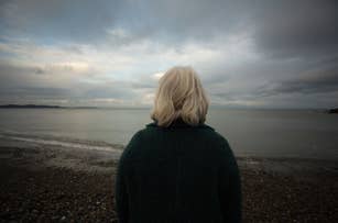 A person with short white hair stands on a pebbled beach, facing the calm sea under a cloudy sky