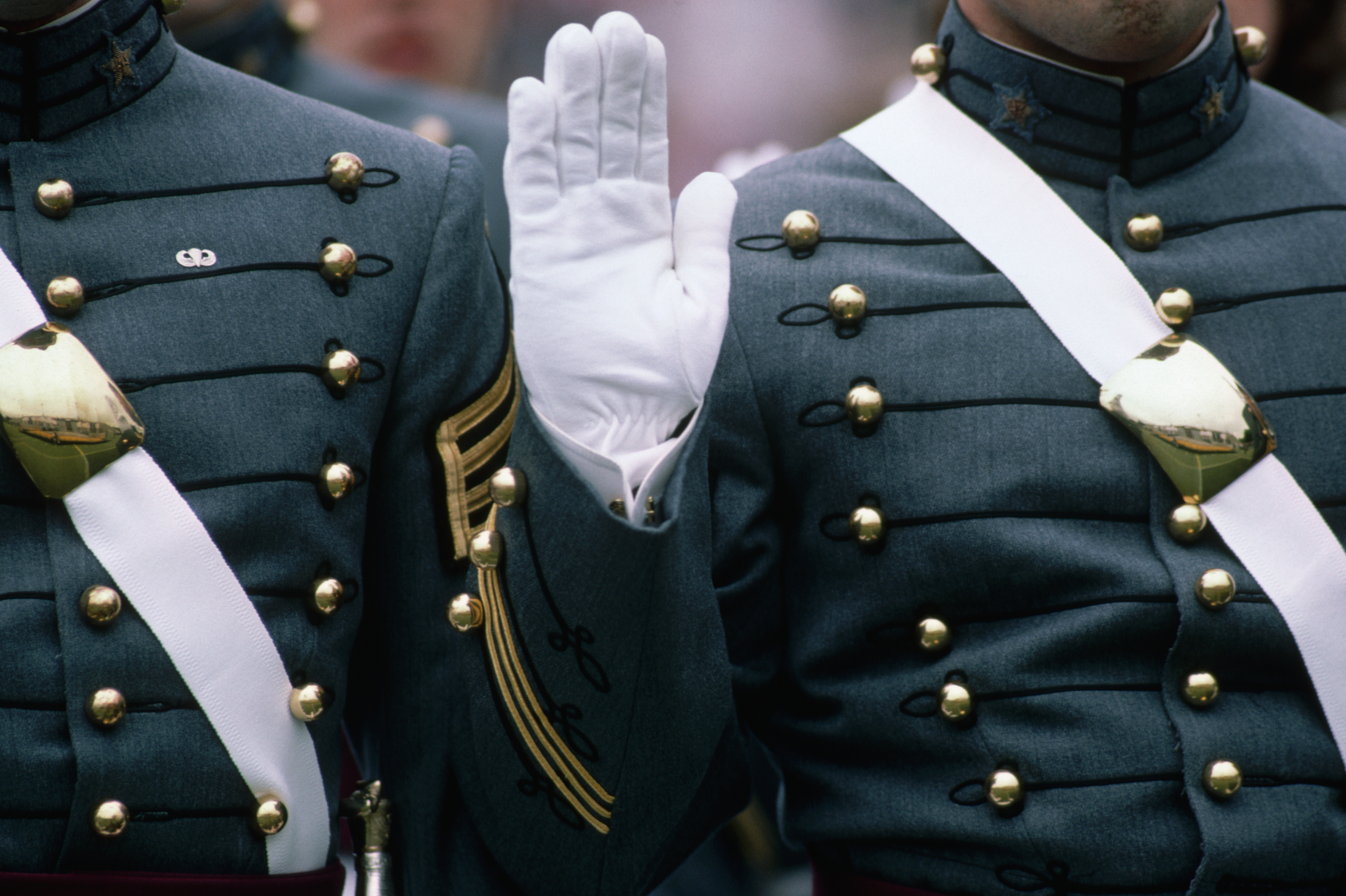 Cadets in ornate military uniforms with white gloves and gold buttons stand at attention, one holding up a hand in salute