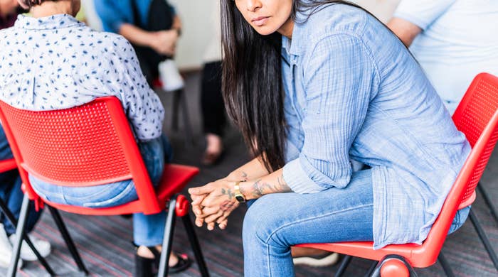 A group of people sit in a circle on red chairs during a discussion. A woman in a blue shirt is seated slightly apart, looking pensive