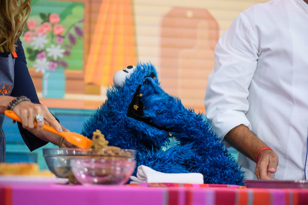 Cookie Monster, woman in chef's coat holding cookie dough, standing next to a person in a white shirt. Cookie Monster is eating cookies. The scene is in a colorful kitchen