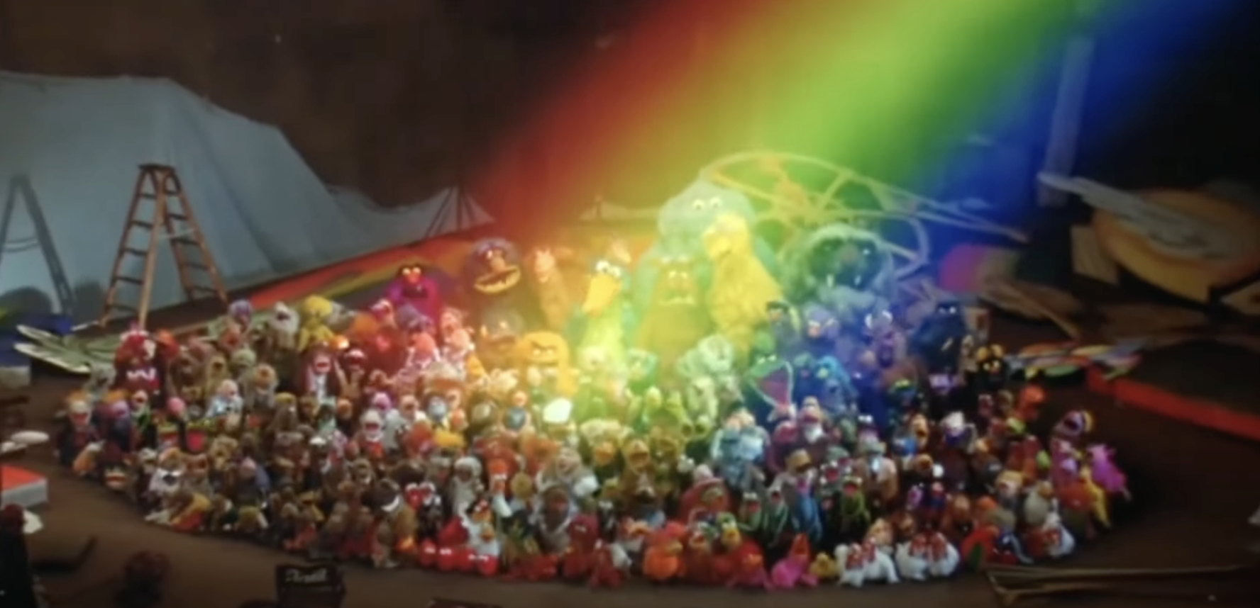 A large group of Muppet characters, including Big Bird and Kermit the Frog, are gathered in a studio setting under a colorful rainbow light
