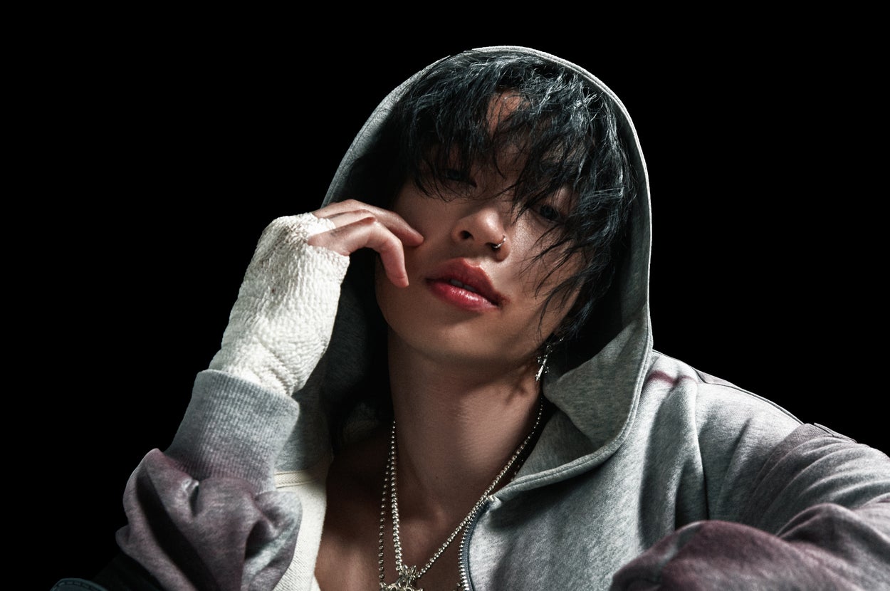 Musician in a hoodie and bandaged hands, with jewelry and an open jacket, sits thoughtfully with head tilted and hand on face