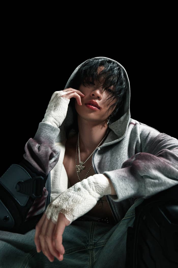Musician in a hoodie and bandaged hands, with jewelry and an open jacket, sits thoughtfully with head tilted and hand on face