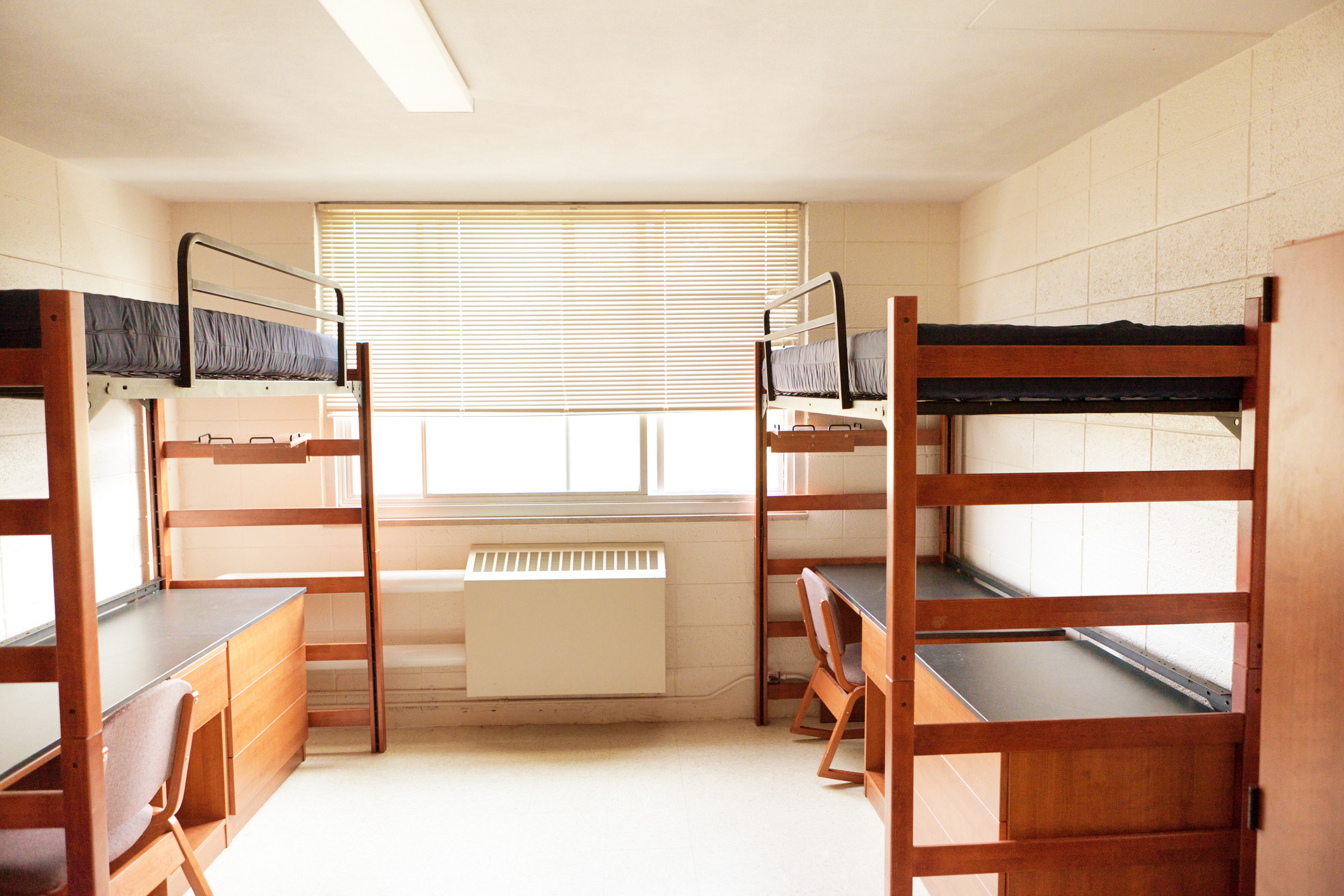 A minimalist dorm room featuring two wooden bunk beds, each with desks and chairs underneath, and a window with blinds in the background