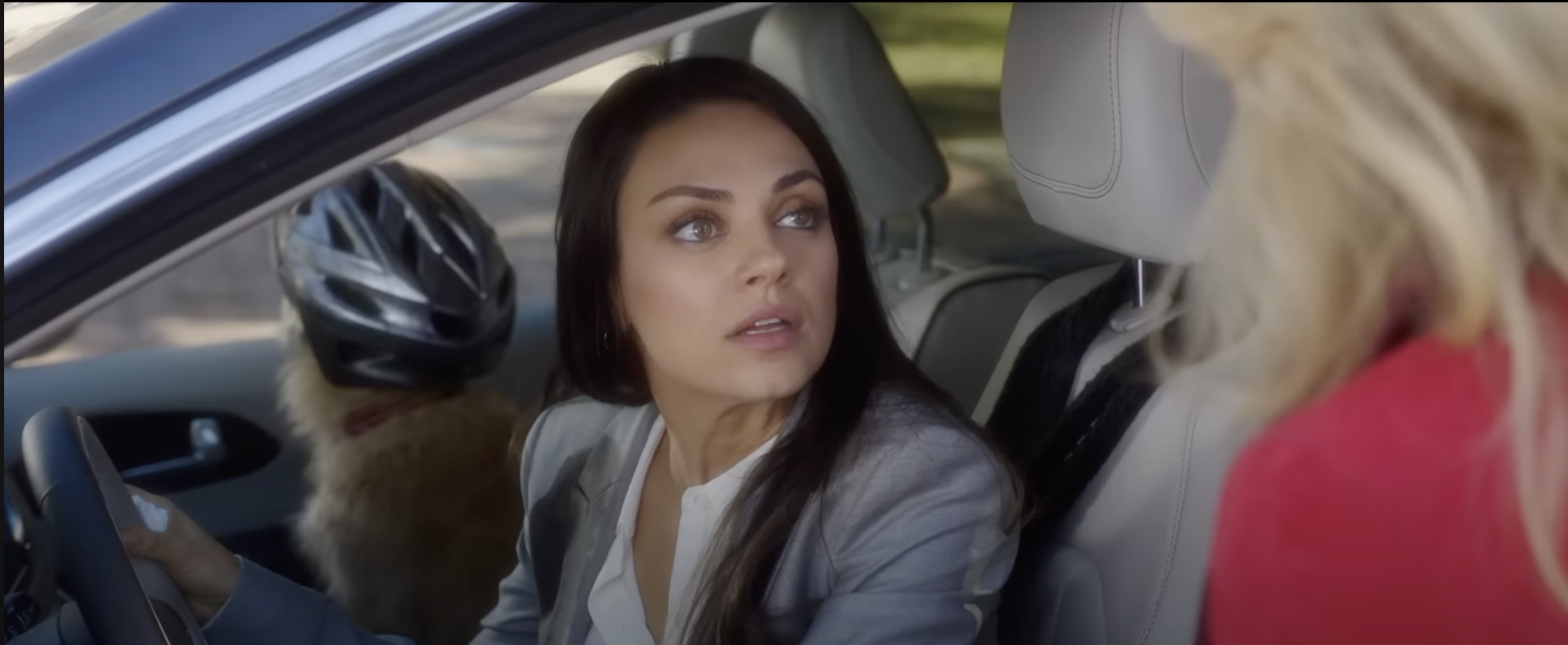 Mila Kunis sits in a car, wearing a white shirt and a gray blazer, speaking with an unseen woman outside the car. A dog with a bike helmet is in the backseat