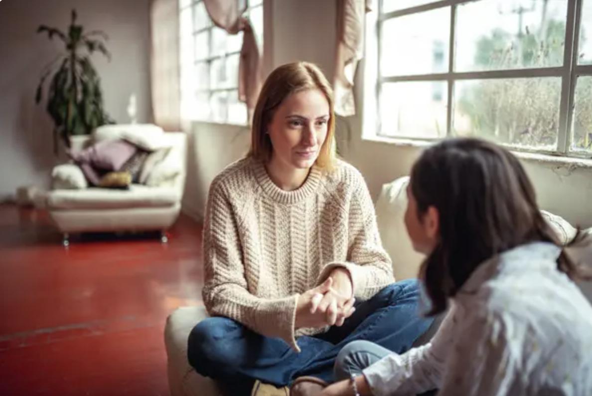 Two women sit on a couch by large windows, engaging in a focused conversation. One woman is wearing a sweater, while the other wears a collared shirt