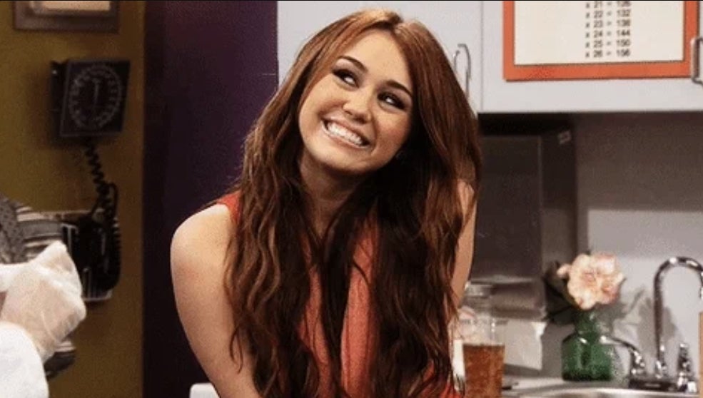Miley Cyrus smiling while sitting in a room with a whiteboard and flowers in the background