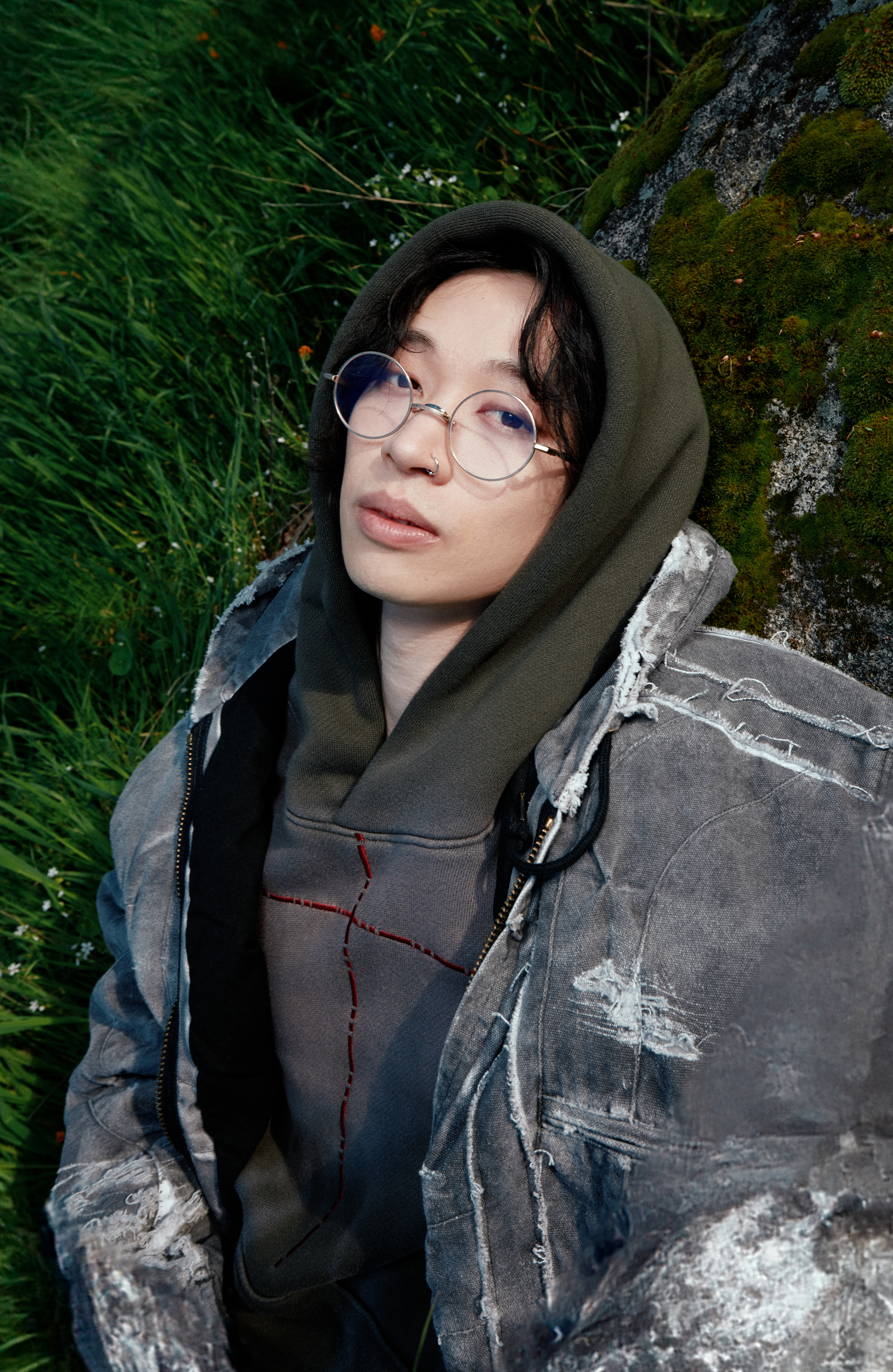 A person with glasses, wearing a distressed jacket and hoodie, reclines on grass beside a moss-covered rock
