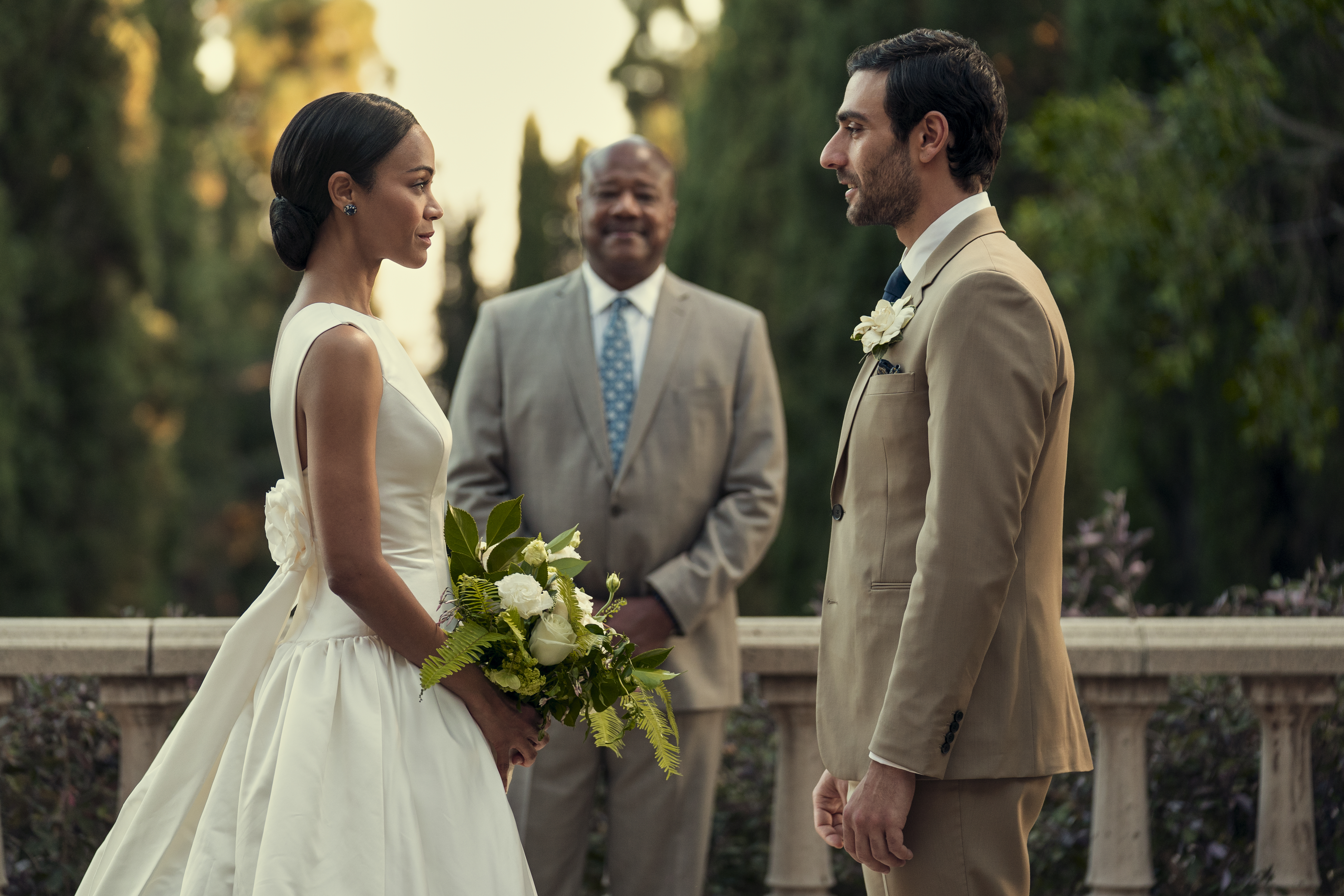 Zoe Saldana and Eugenio Mastrandrea facing each other in wedding attire, with Keith David smiling behind them, in an outdoor setting in a scene from "From Scratch."