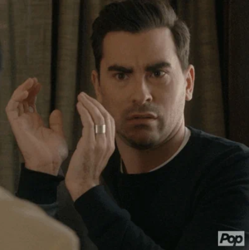 Dan Levy looking surprised, with his hands raised near his face, wearing a black sweater in a scene from "Schitt's Creek."