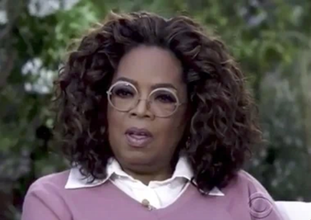 Oprah Winfrey, wearing glasses, a white shirt, and a pink sweater, speaking during an outdoor interview